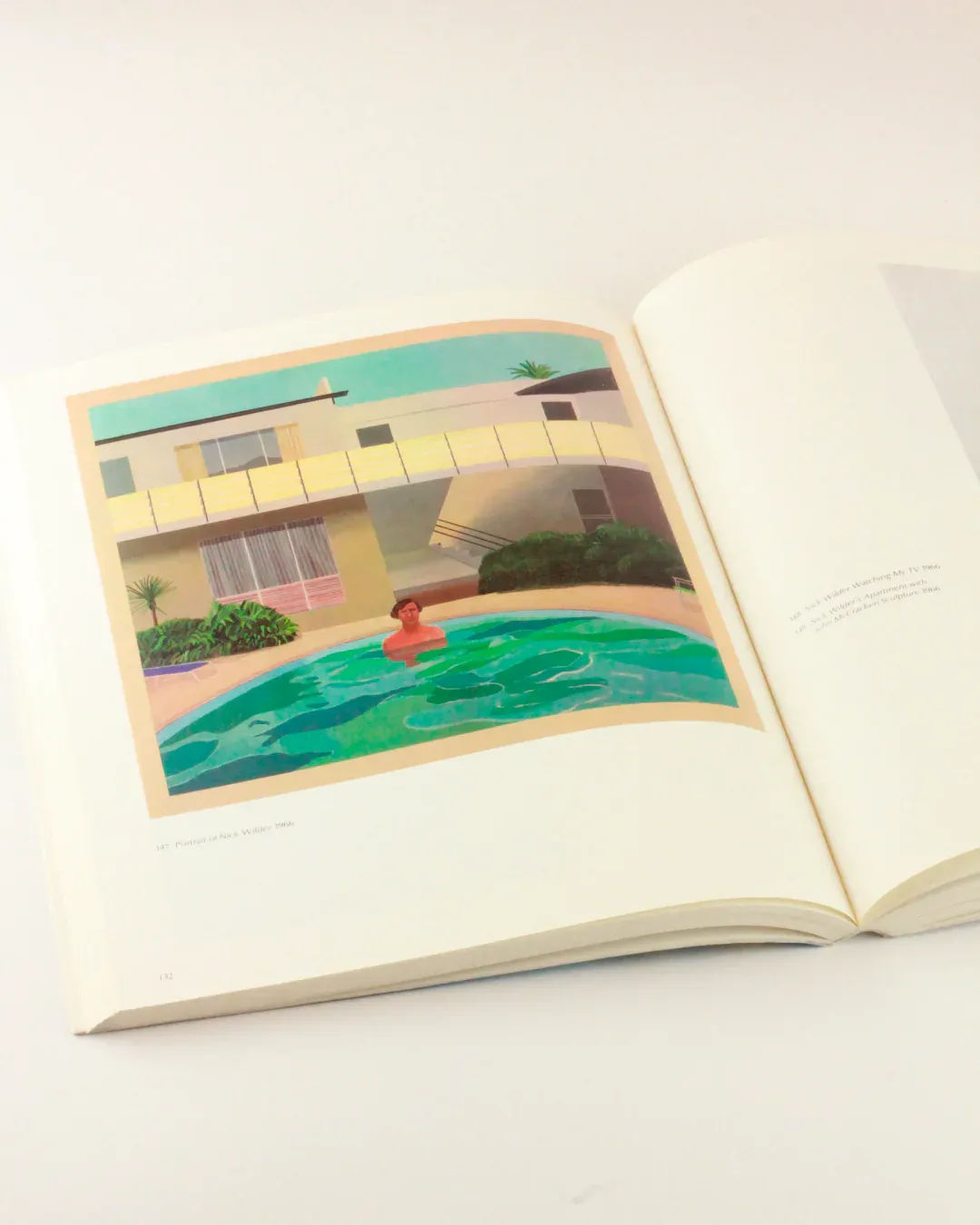 An open book displaying an interior page featuring a painting by David Hockney of a person swimming in a pool located in front of a modern house with large windows and a balcony. The surrounding greenery creates a serene scene, evoking the essence of arte británico. The background is white. This is David Hockney 'My Early Years' from Boga Avante Shop.