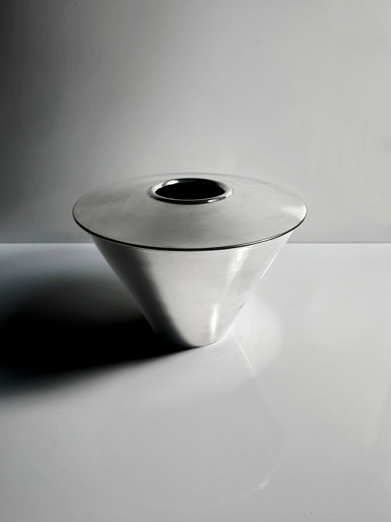A sleek, polished metal cone-shaped Heavy Silver-Plated Vase from Fink Germany by Dodo Vintage features a circular opening on top. Set against a minimalist gray and white background, the lighting casts subtle shadows highlighting its smooth surface.