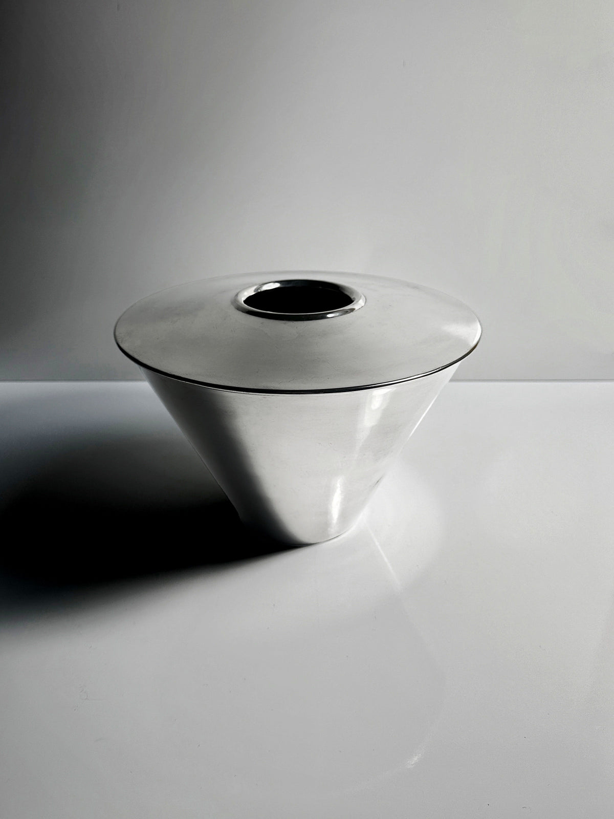 A sleek, polished metal cone-shaped Heavy Silver-Plated Vase from Fink Germany by Dodo Vintage features a circular opening on top. Set against a minimalist gray and white background, the lighting casts subtle shadows highlighting its smooth surface.