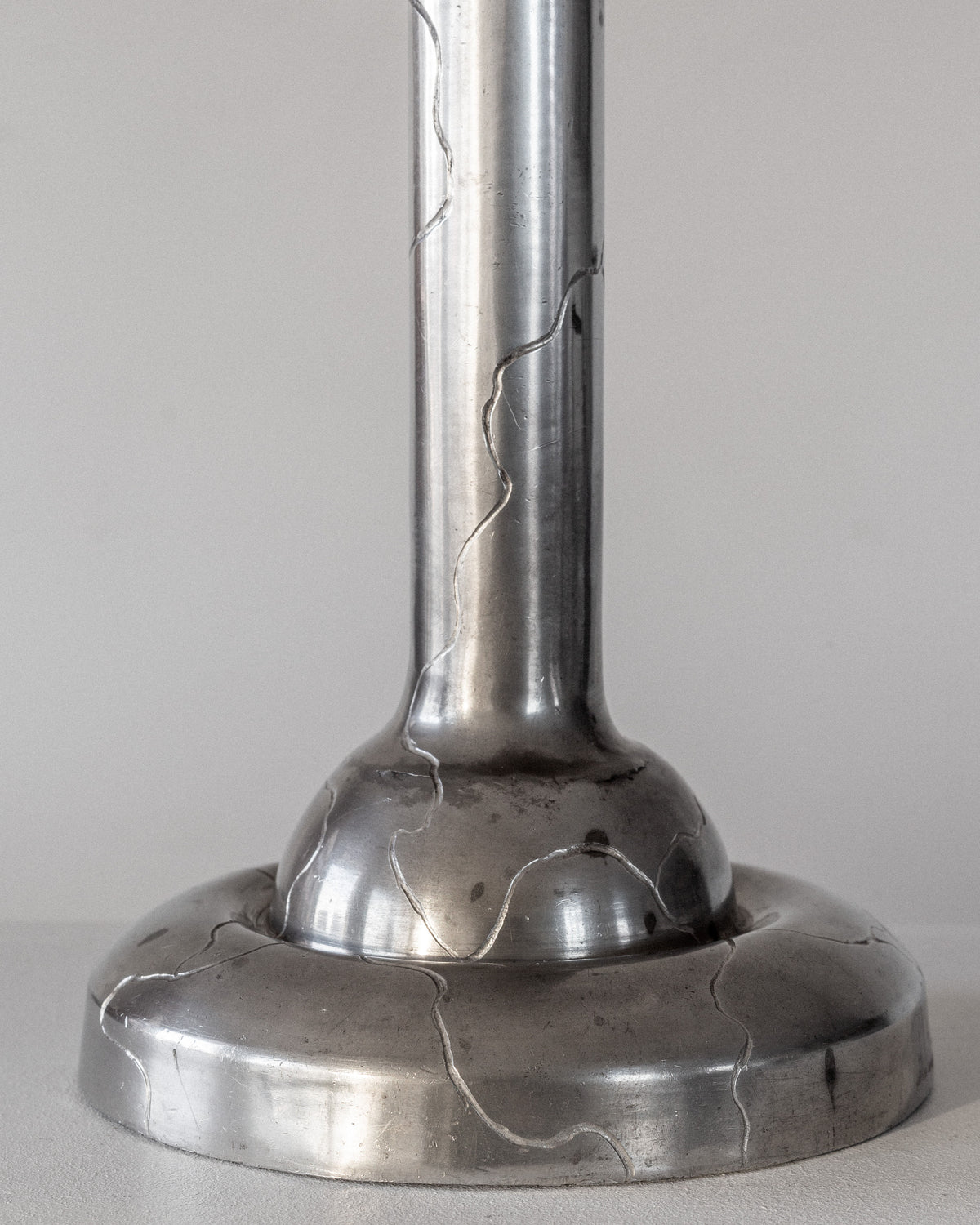 A close-up of the Spigel Art Deco Pewter Lamp from 1920s Sweden, featuring a cylindrical design and circular base with visible gold-filled cracks as kintsugi repair. The background is plain and neutral-colored.