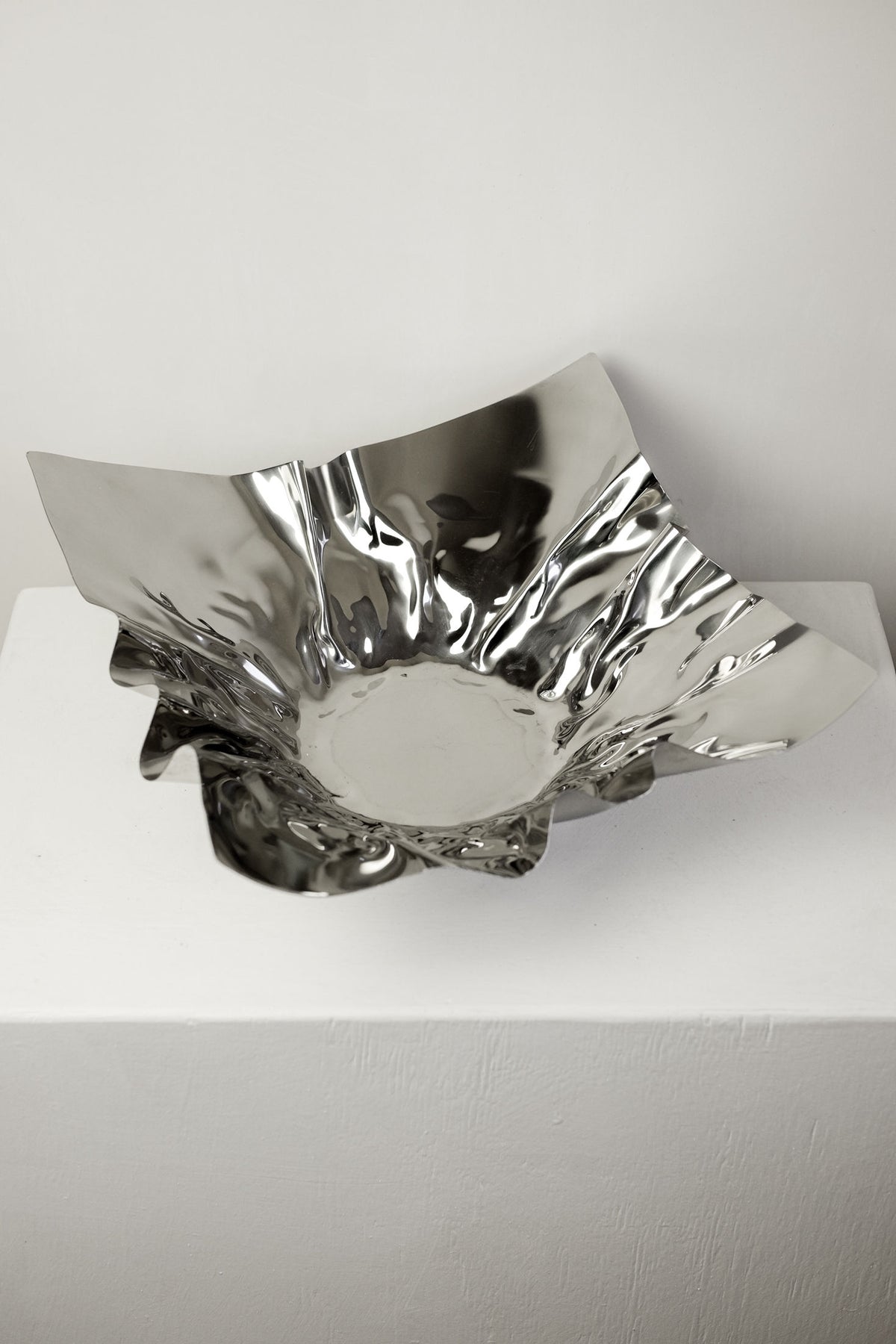 The Extra Large Folded Chrome Bowl by septembre studios features jagged, uneven edges and a crumpled, reflective surface, adding vintage charm to any space when displayed on a white pedestal against a light gray background.
