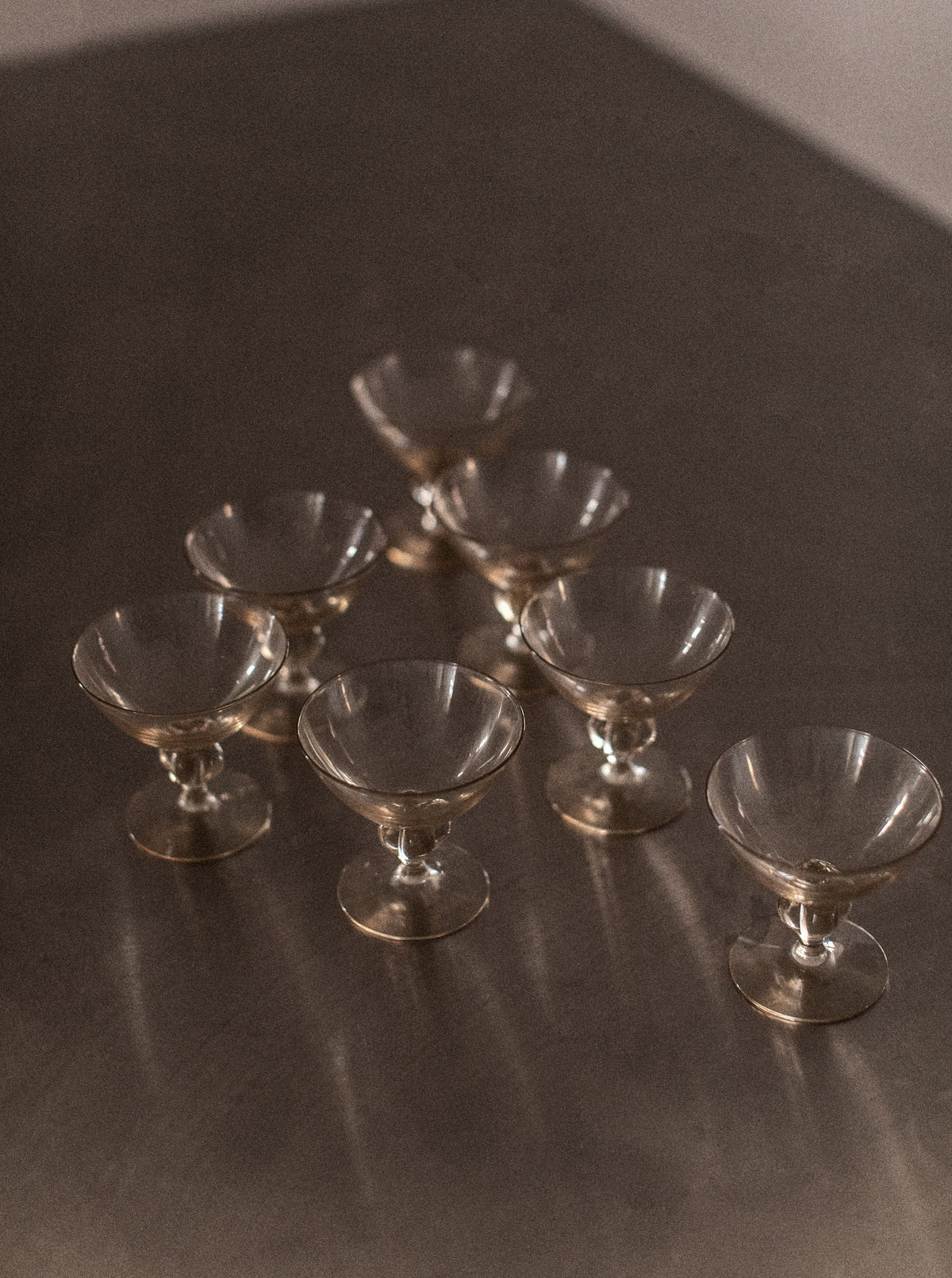 A group of seven empty *Set of 7 Art Deco Glass* cups from *Out For Lunch*, arranged in a pyramid-like formation, sits on a reflective surface. The lighting is soft and creates subtle reflections on the surface, emphasizing the delicate transparency and contours of the glasses, elevating the drinking experience with an air of sophistication.
