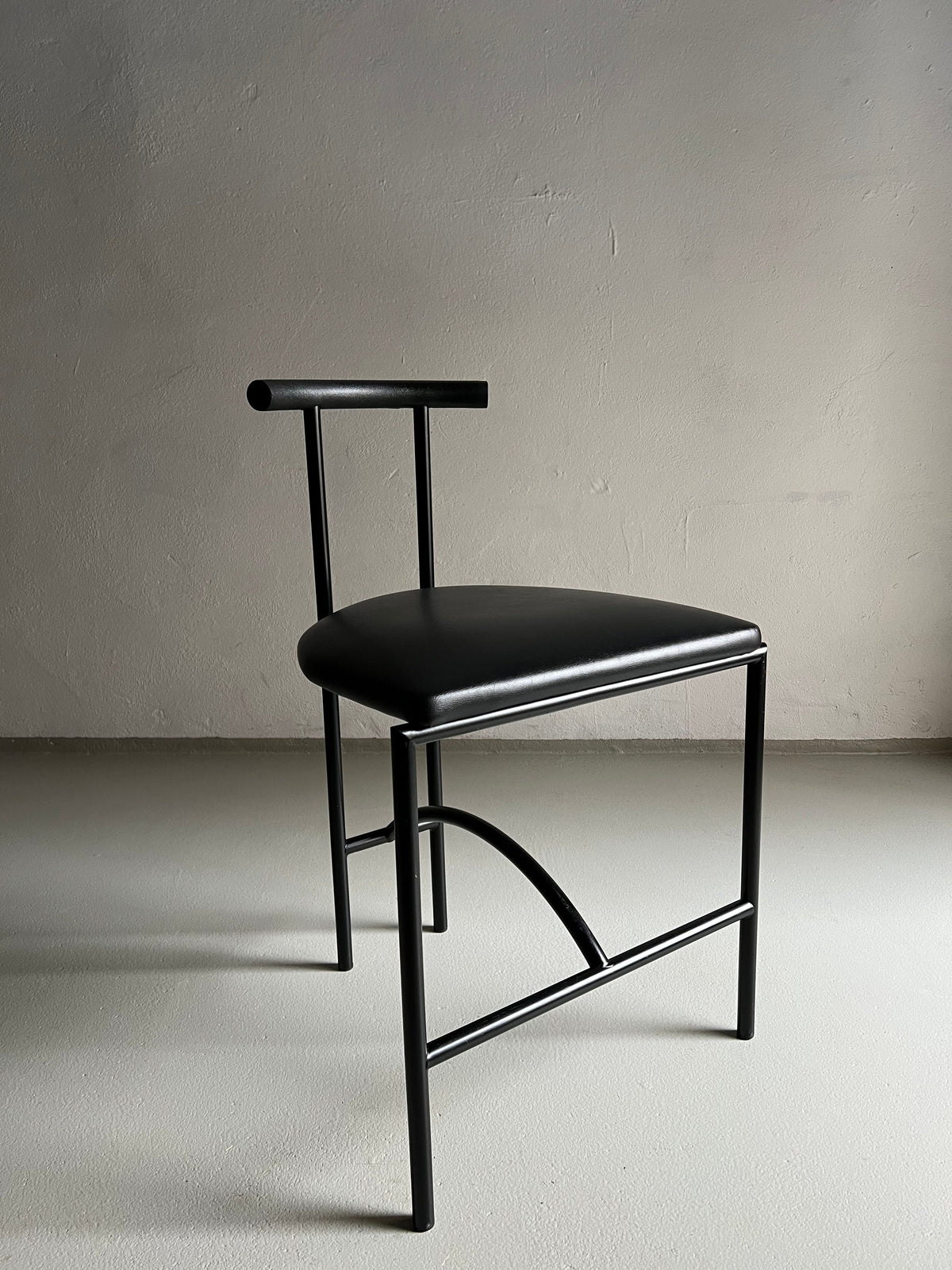 A minimalist black metal chair with a unique geometric design, featuring a T-shaped backrest, simple faux leather upholstery seat cushion, and curved support bars underneath. The chair is placed against a plain light-colored wall and sits on a smooth floor. Product Name: 4 Postmodern "Tokyo" Chairs by Rodney Kinsman | Italy | 1980s Brand Name: Veter Vintage.