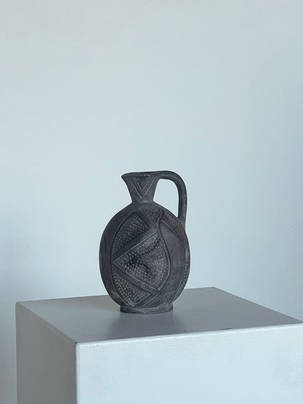 The "Jug with Hand Markings" by Porter Studio, a dark handmade piece with intricate designs and a handle, is showcased on a white pedestal against soft gray tones. This artisan pottery jug displays symmetrical patterns etched across its surface.