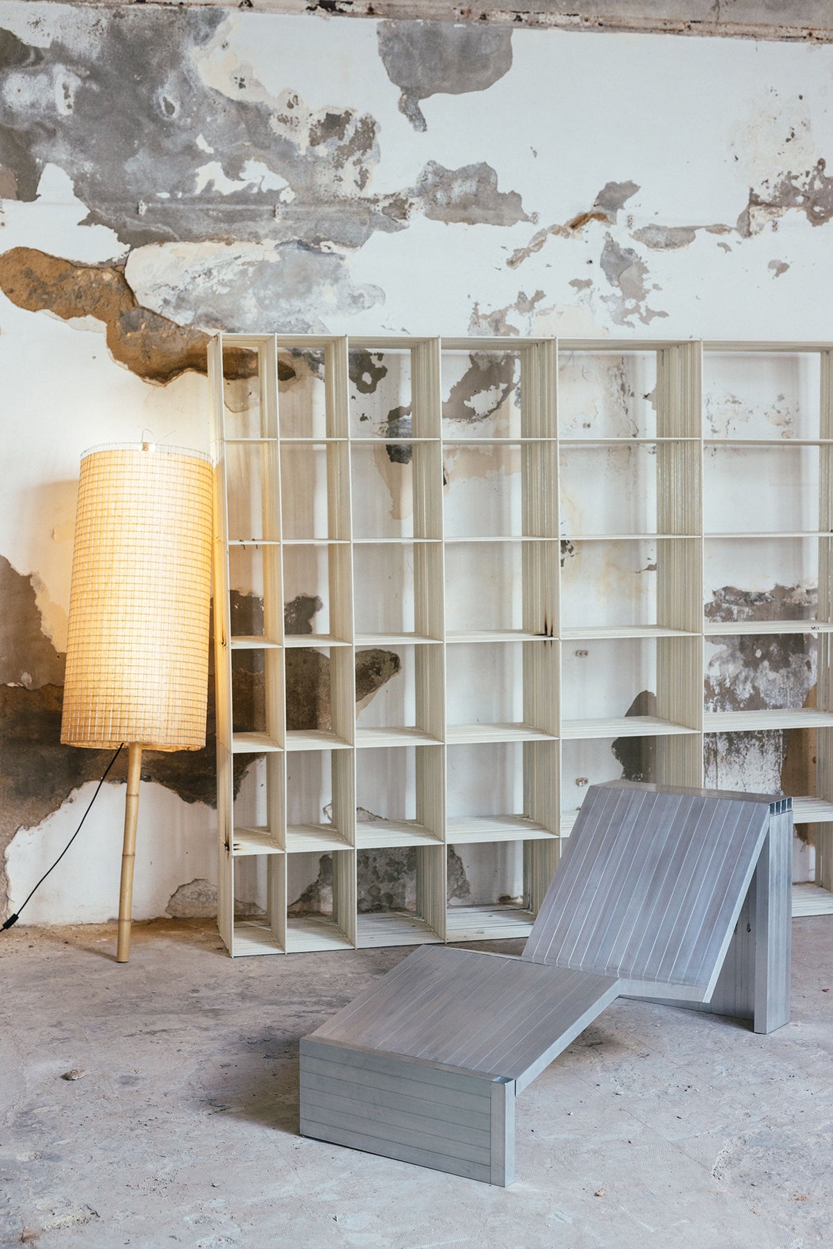 An Aluminum Lounge Chair by Studio HAOS sits beside empty wooden shelves and a tall floor lamp with a woven shade, all set against a weathered wall in an industrial-style room with modern accents.