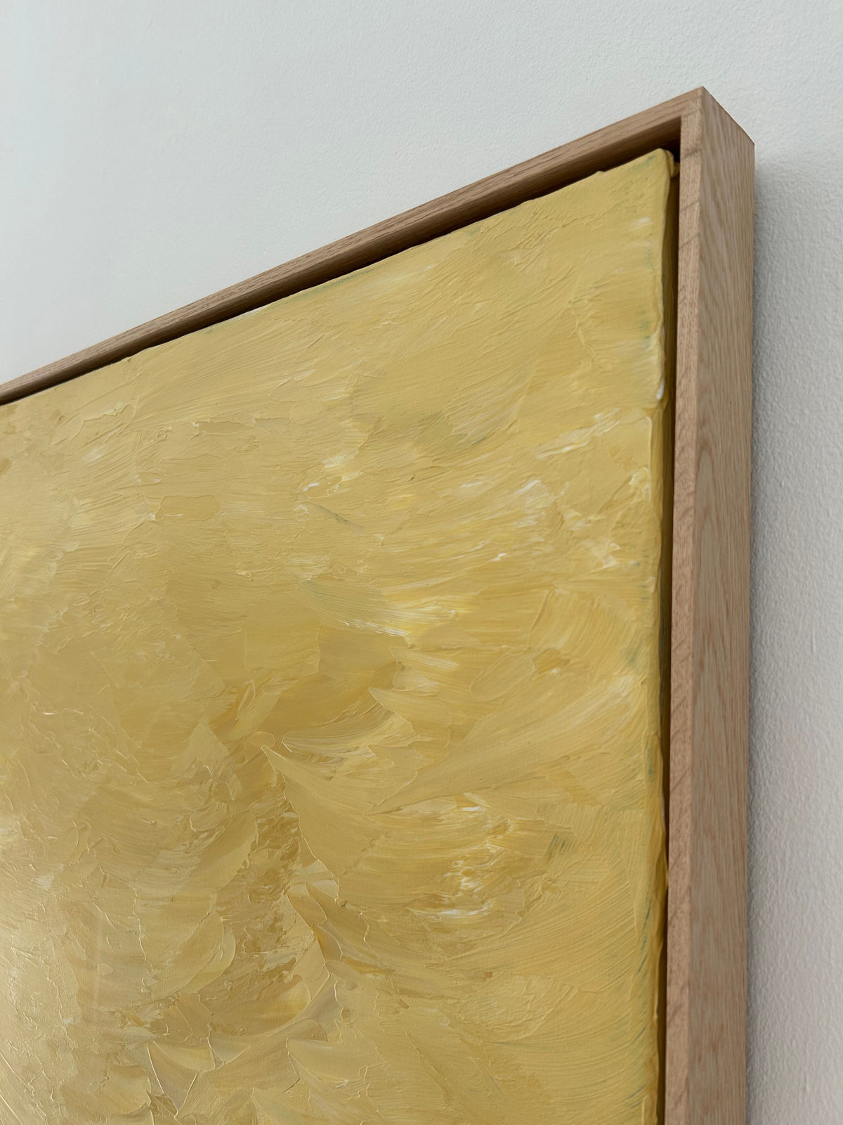 A close-up view of a framed painting featuring a textured, abstract design in varying shades of yellow. This mixed media piece from the Mattias Storm Solar Mirage - Mixed media on canvas series is encased in an oak frame and mounted on a white wall.