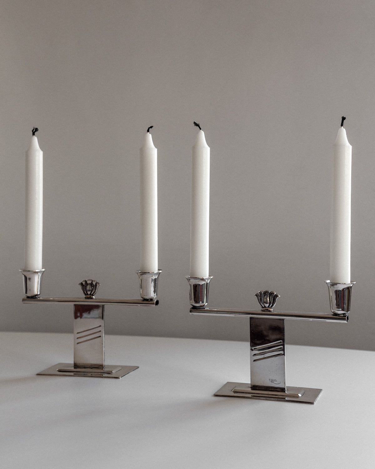 The pair of Silver-Plated Art Deco Candle Holders by Spigel elegantly hold two tall, unlit white candles each, placed on a white surface against a neutral backdrop. These candleholders exhibit sleek Scandinavian craftsmanship.