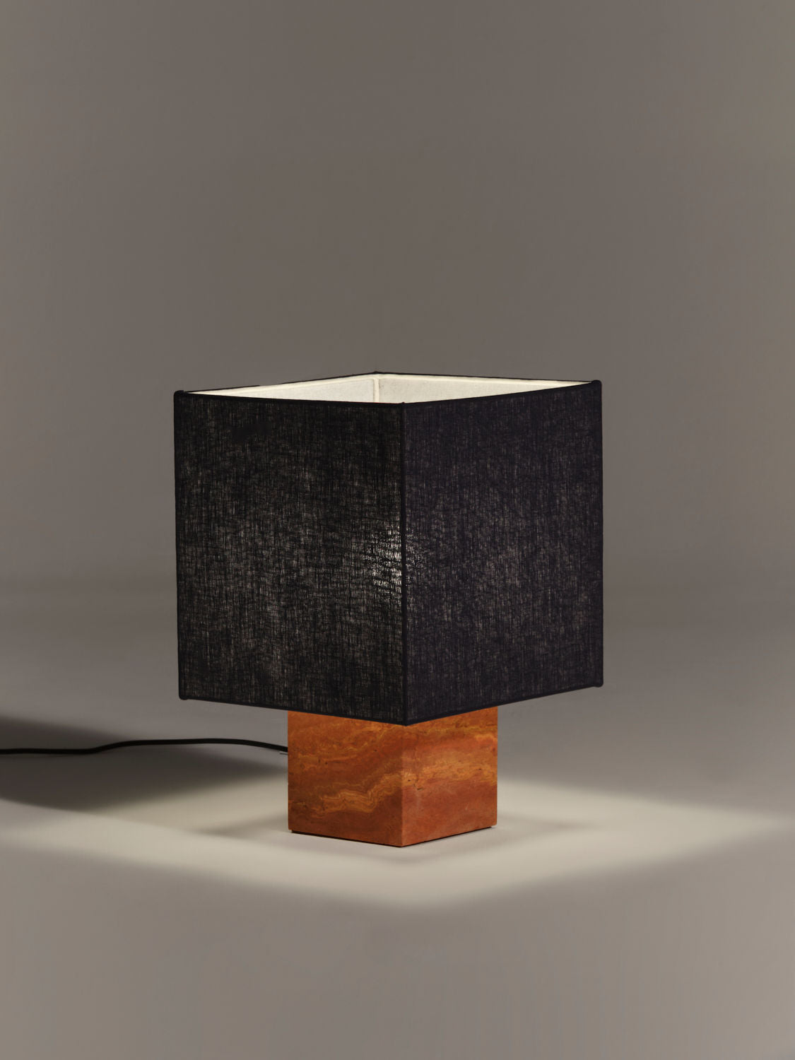 A minimalist table lamp with a black square shade and a wooden square base, crafted from natural materials, set against a plain grey background. The Nassi Lamps Roma I Squared Red Travertine is illuminated, casting light onto the surface around it.