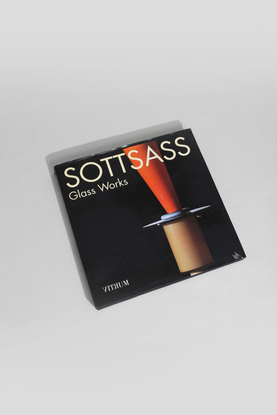 The Ettore Sottsass, Glass Works Book by Villa features a square black cover with a red and orange glass sculpture and highlights Sottsass’s stunning glass works. The book is shown resting on a light gray surface.