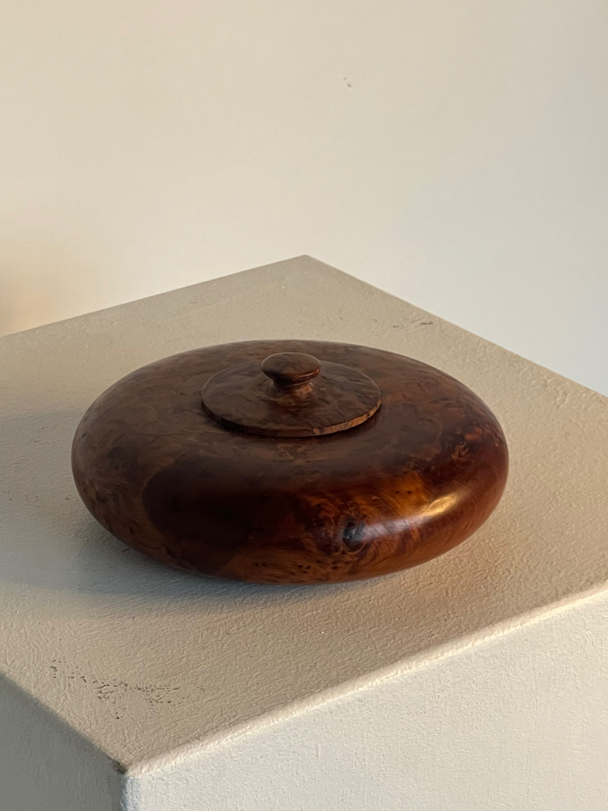 The Walnut Burl Shallow Bowl With Lid by Porter Studio is a beautifully crafted round dish featuring a small knob on top. Made from rich brown walnut burl, it showcases a smooth, glossy finish with visible grain patterns. Elegantly displayed on a white pedestal, the shallow bowl sits against a simple beige background.
