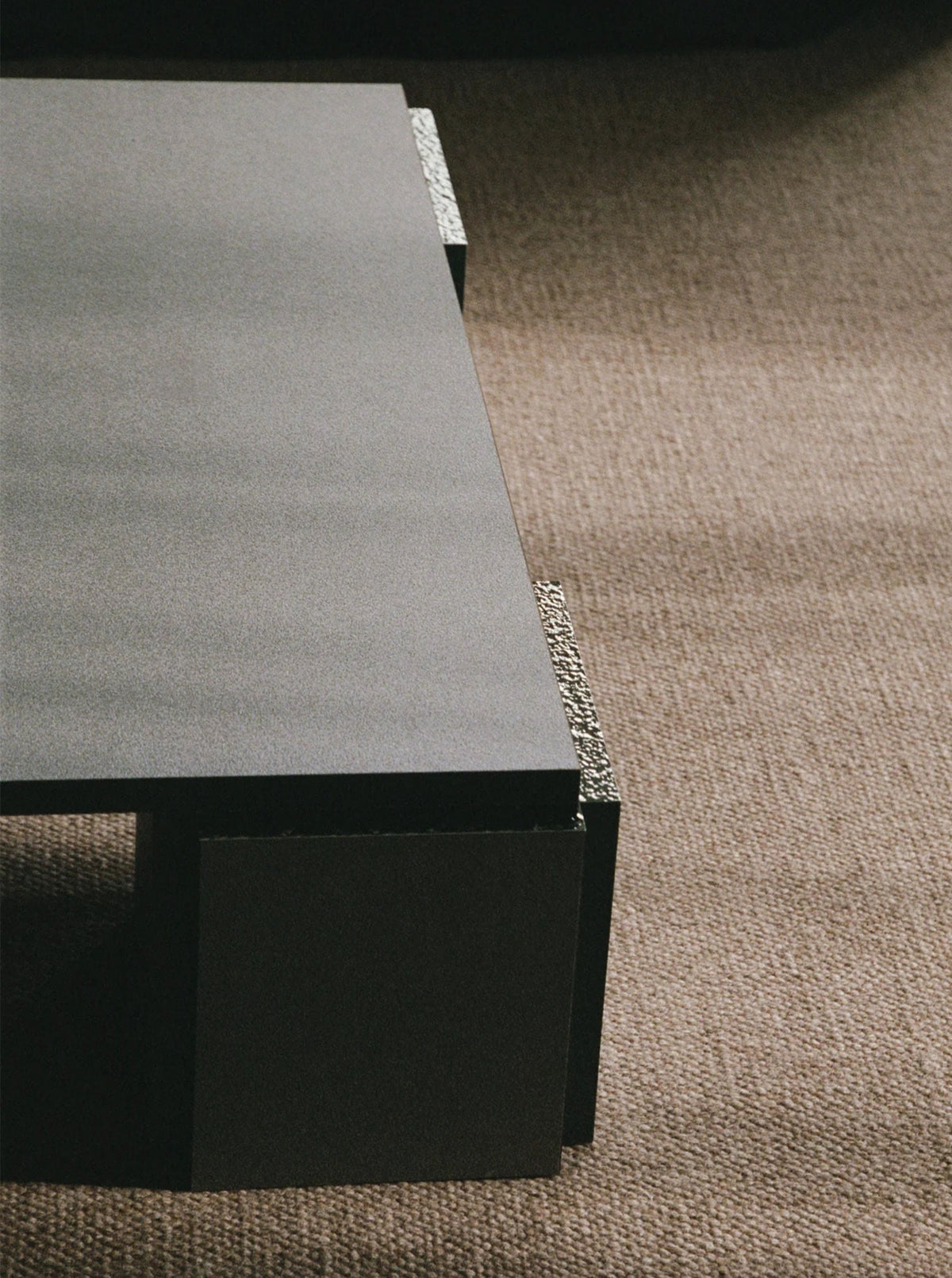 A low-profile, minimalist "Mara" Coffee Table – Night Noise by TEGET features a textured edge and sits on a woven brown carpet. The sharp angles and dark color of this piece of laminated furniture create a striking contrast with the soft, neutral-toned carpeting.