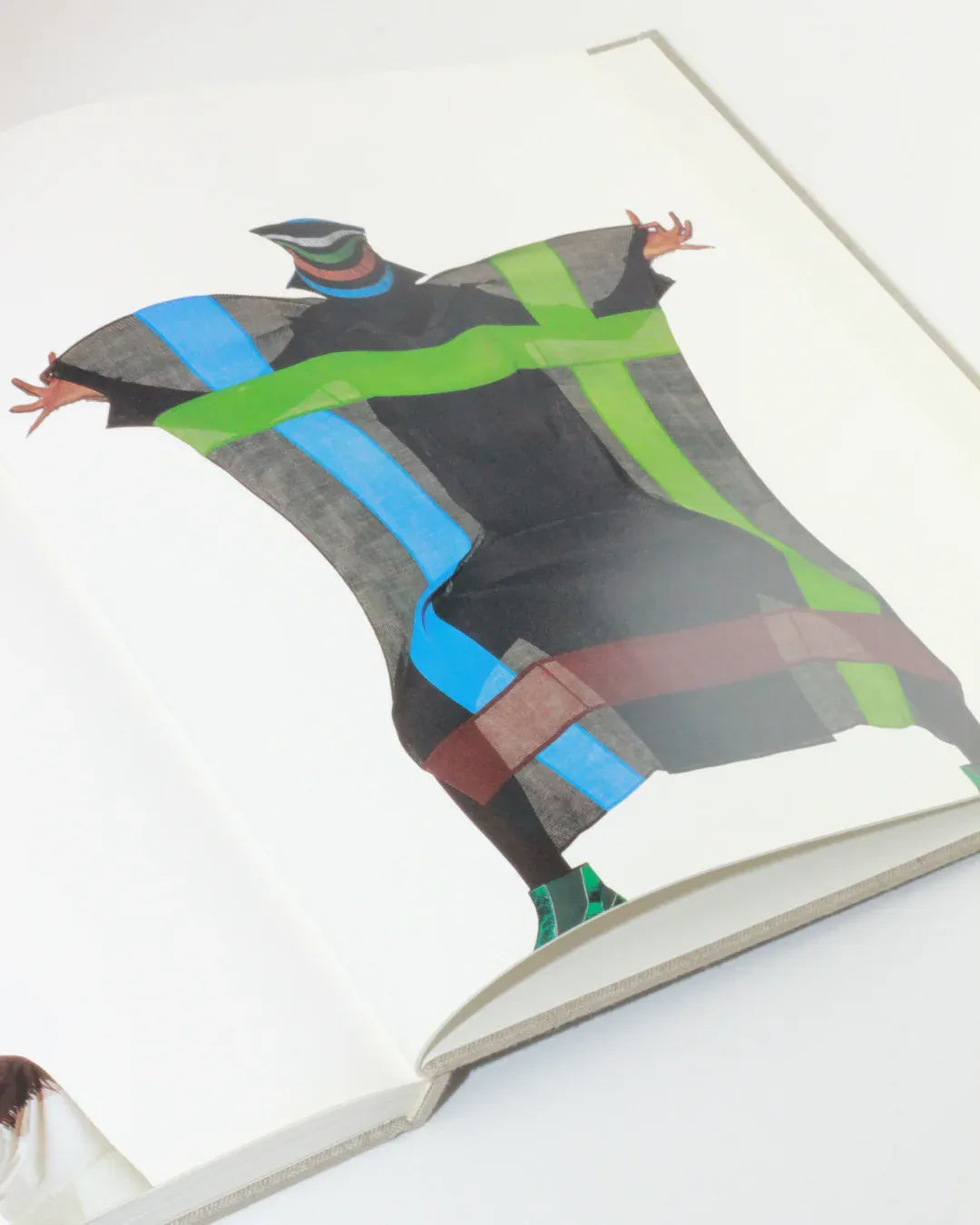 An open book from Boga Avante Shop titled "Irving Penn regards the work of Issey Miyake" is displayed on a white surface, featuring an illustration of a person in a dynamic pose. The figure is adorned in a long, colorful coat with bold, wide stripes of black, green, blue, and brown that mostly obscure their face and legs, evocative of Irving Penn's striking fashion photography.