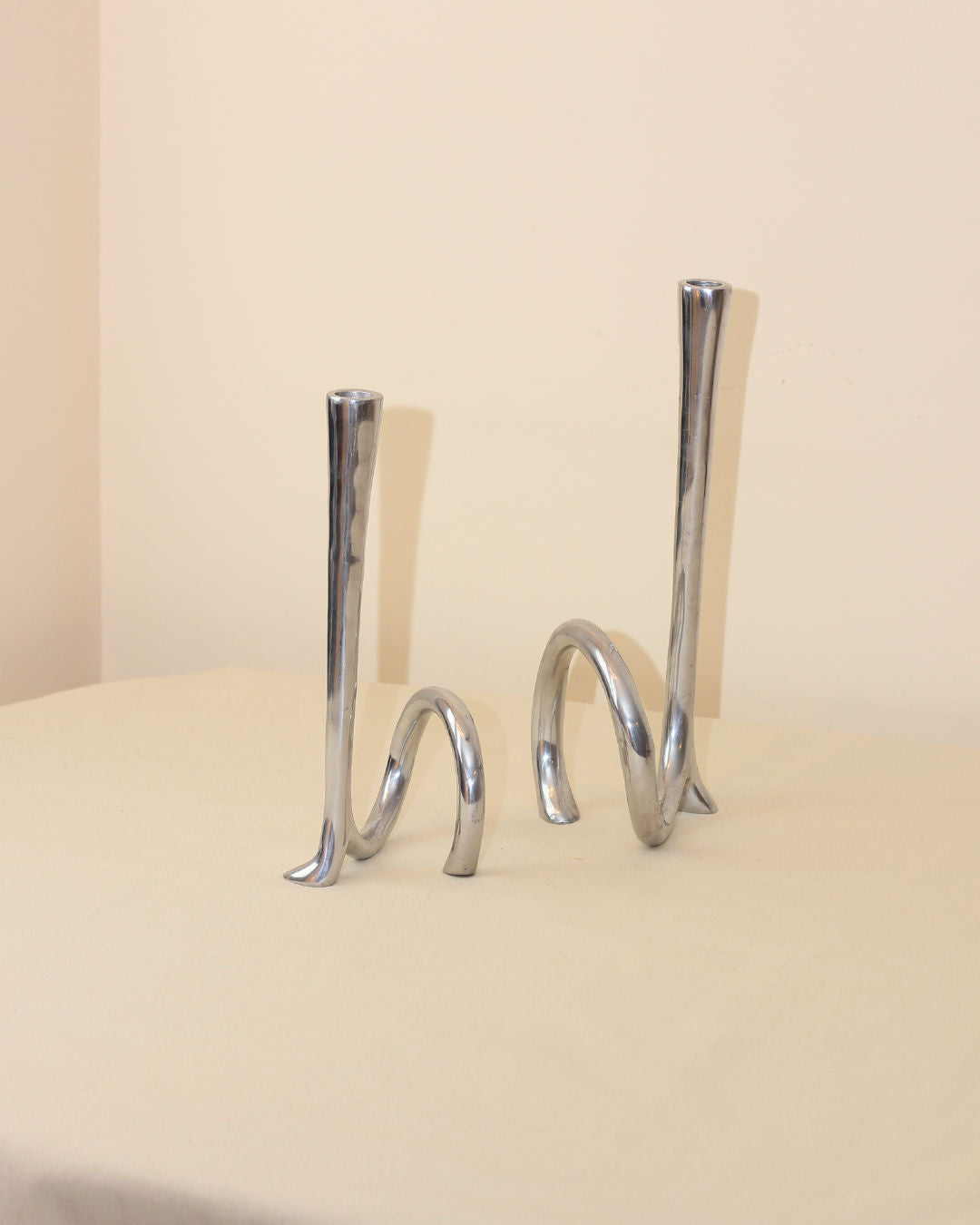 Two abstract Cast Aluminum Candleholders by Boga Avante Shop feature curved, twisted bases inspired by 1980s candelabra, ideal for modern minimalist decor on a beige surface against a plain, light-colored wall.