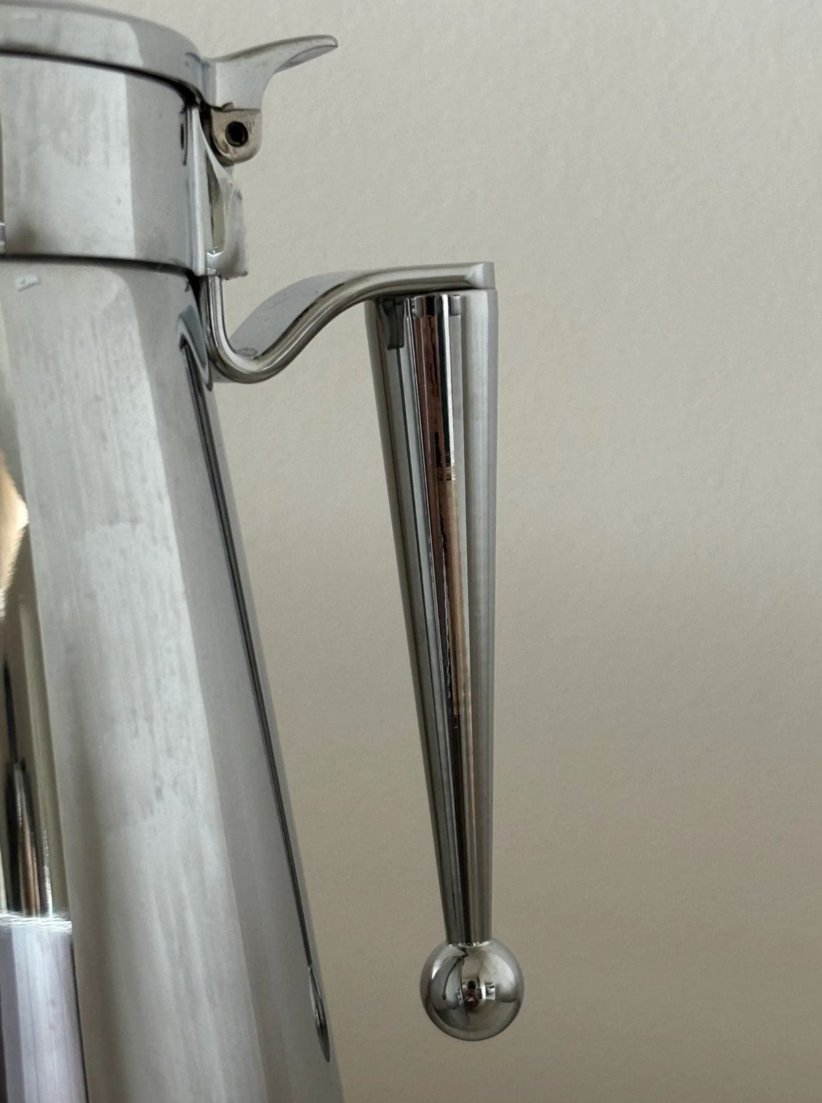 Close-up of a silver metallic coffee pot handle against a beige background. The handle, part of a CU-RA-TED Steel Kettle “Erhard Germany,” is sleek with a conical shape that narrows towards the bottom and ends in a spherical knob. The pot's polished surface reflects light, showing its impeccable condition.