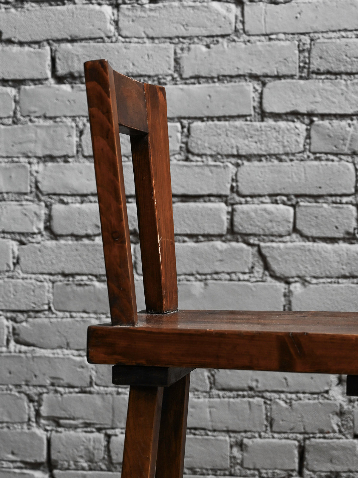 A close-up side view of a Studio OSKLO Set of 8 Geometric Wooden Chairs 40s shows a rustic oak dining chair with a simple, angular dark brown backrest and seat, set against a light gray brick wall.
