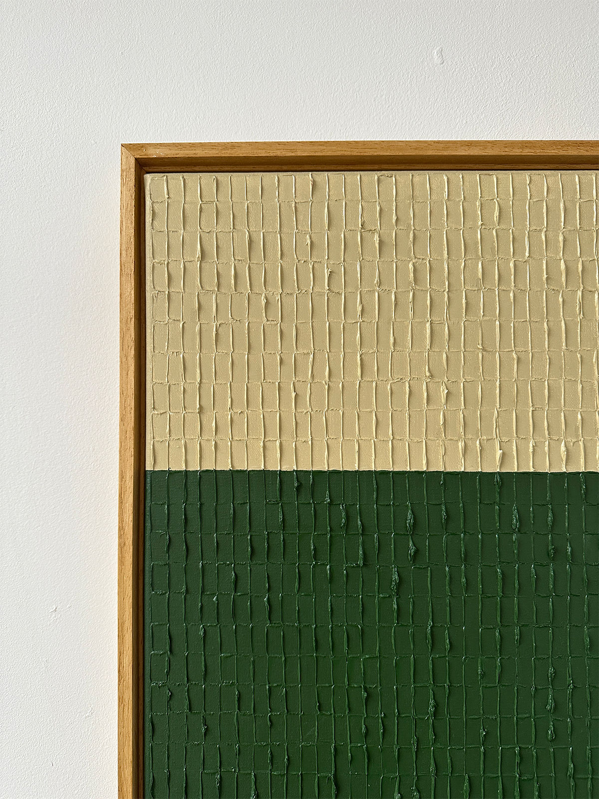 The "Buff Verte" by Roi Elmaliah is a textured abstract painting created with oil on canvas, showcasing a unique design divided into two sections: the top half in beige and the bottom half in dark green. It features an intricate grid pattern and is elegantly framed with a wooden border, all displayed against a simple, light-colored wall. This piece draws inspiration from Amsterdam's vibrant artistry.