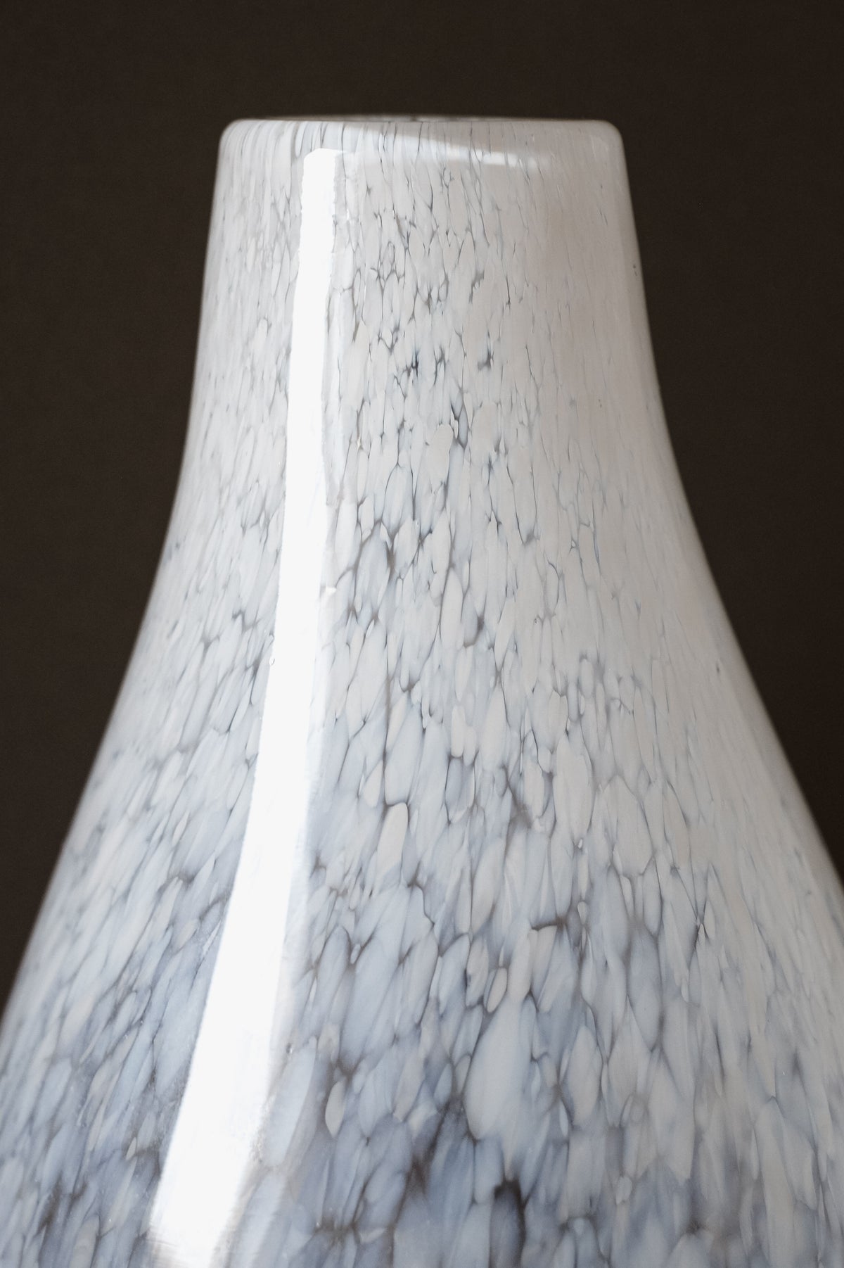 The White Vase Tortoise Glass by septembre studios exudes a glossy, vintage charm with its surface of gray and white speckles that create a marble-like effect, standing elegantly against a dark background.