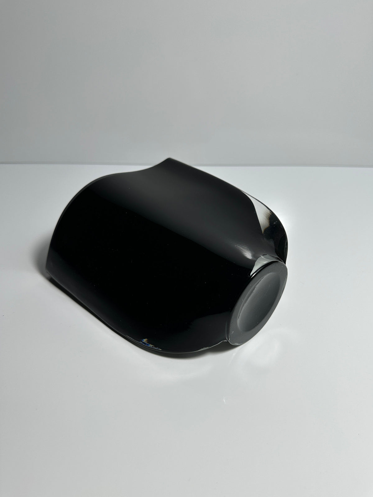 A sleek, black, curved object sits on a white surface against a neutral background. Resembling the modern and abstract design of the Black Glass Vase by Kaj Franck for Dodo Vintage, it boasts a glossy finish that enhances its sophisticated allure.
