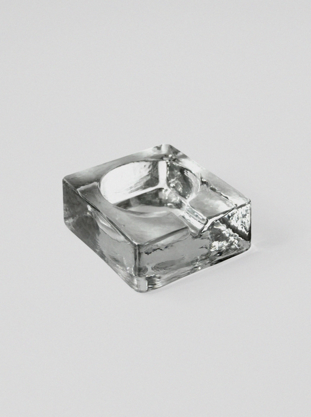 The Brutalist Massive Glass Ashtray Bowl by Vintage Stories is a clear, square Scandinavian piece in a subtle 70s brutalist style with a single cigarette groove, set on a plain light gray background.