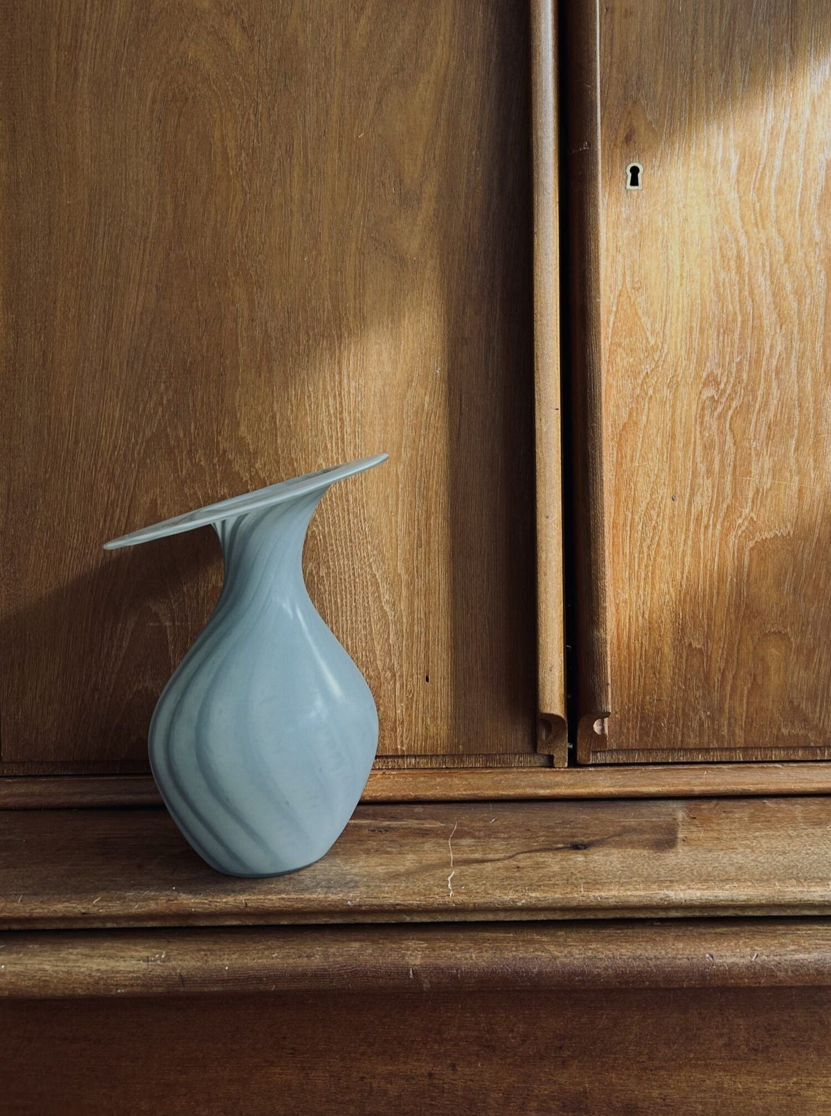 A Vintage Vase Peill & Puztler by Flock Studio, in a pale blue with a distinctive shape and flat top, rests on a wooden surface. The background features wooden panels, with sunlight casting its glow to create a warm, rustic atmosphere that exudes timeless elegance.