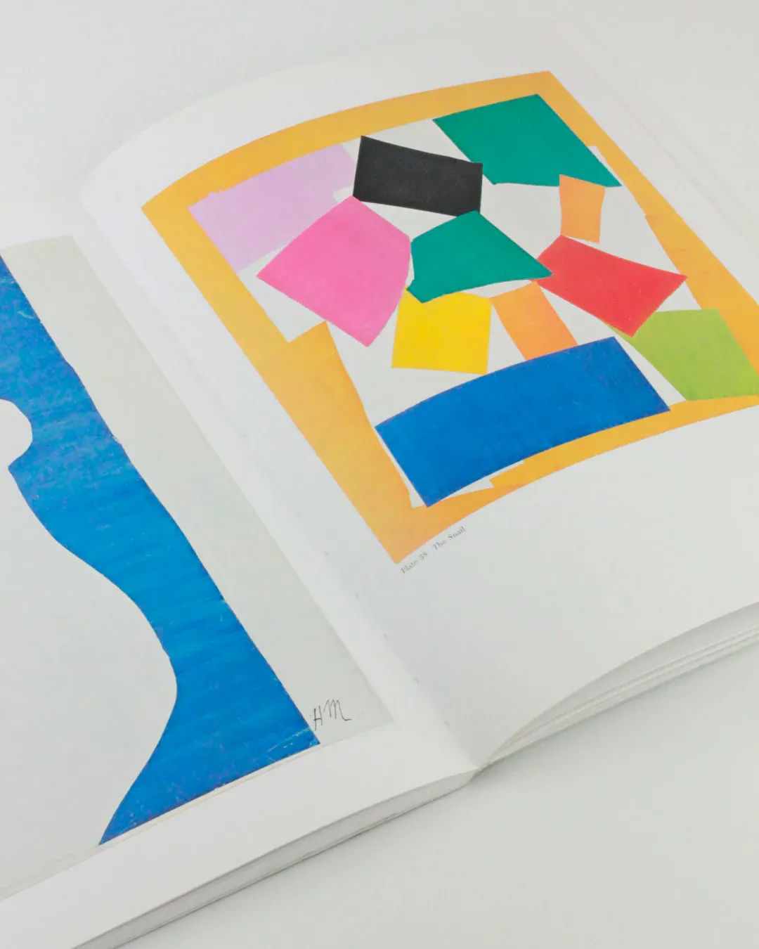 An open art book from Boga Avante Shop titled "The Cut Outs of: Henri Matisse" displaying two pages. The left page features an abstract blue and white design, reminiscent of Henri Matisse's mature cut-outs, while the right page showcases a vibrant collage with pink, black, green, yellow, and blue shapes set against a yellow border.