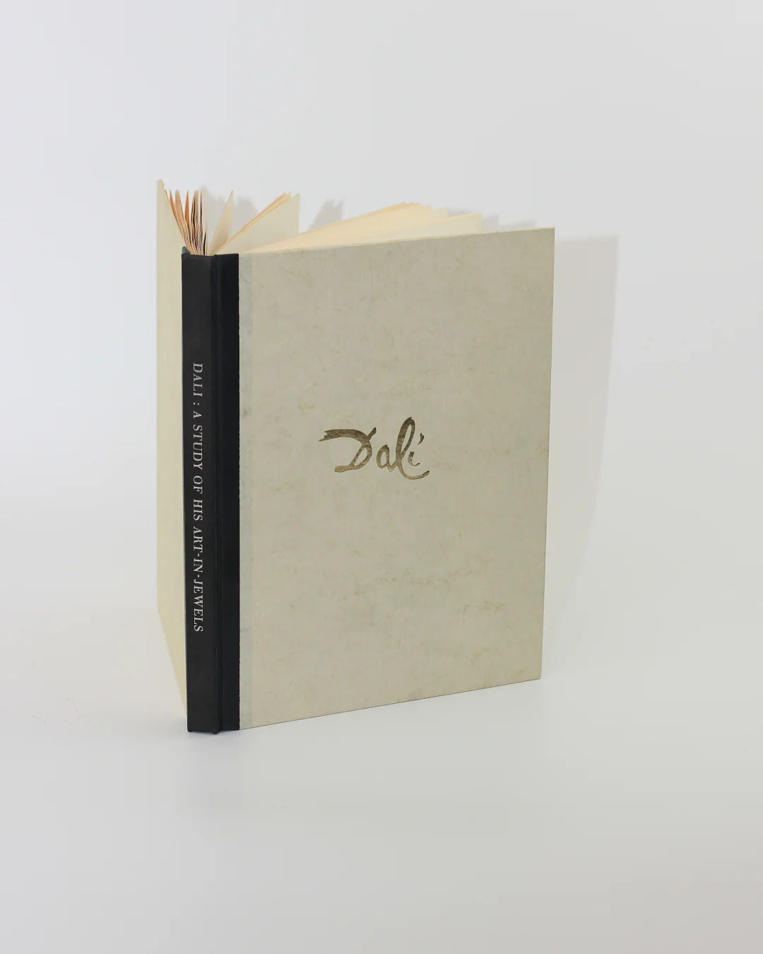 The book "Dalí: A Study of his Art-in-Jewels" from Boga Avante Shop stands against a plain backdrop, showcasing Dalí's surrealist jewelry. Its beige cover has "Dalí" elegantly scripted, with pages gently fanned open to reveal his artistic legacy.
