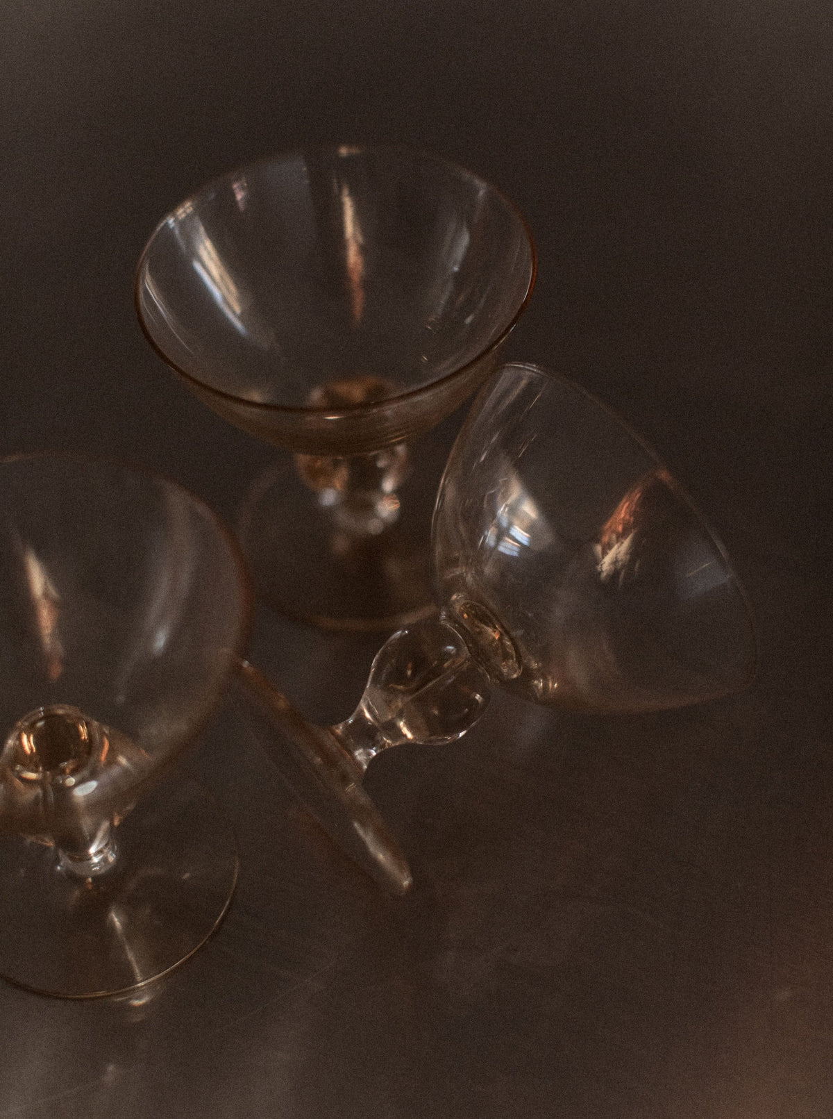Three empty, elegant glasses from the Set of 7 Art Deco Glass collection by Out For Lunch are placed on a reflective surface. Two glasses stand upright while one lies tipped over on its side. The scene exudes sophistication with soft and warm lighting, creating a delicate and moody atmosphere that hints at an intimate drinking experience.