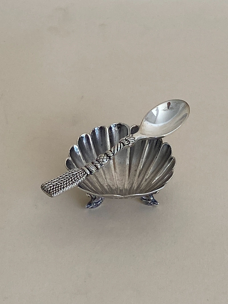 The French Tassel Spoon by Aesthete Label includes a small, ornate silver dish in excellent vintage condition, featuring a scalloped shell design supported by three tiny legs. It pairs perfectly with the matching silver spoon, which boasts a textured handle that rests elegantly across the dish—an exquisite addition to any decorative table setting. The background is plain and light-colored.