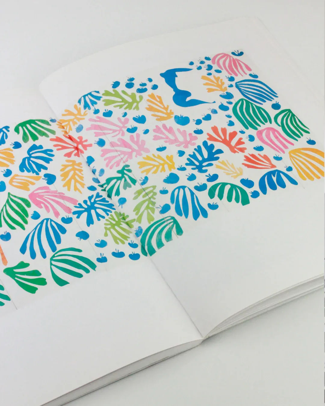The book "The Cut Outs of: Henri Matisse" from Boga Avante Shop lies open on a white surface, showcasing a vibrant abstract design that echoes the style of Henri Matisse's mature cut-outs. The page features an array of leaf shapes in green, blue, pink, and orange, interspersed with small blue dots on a white background, enhancing the vividness of the colors.