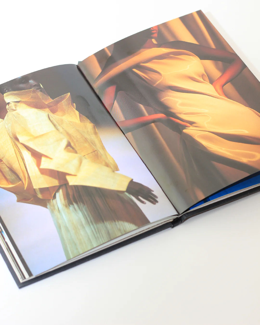 An open Issey Miyake Book from Boga Avante Shop reveals two pages. On the left page, a model wears a sculptural, layered yellow outfit that highlights Issey Miyake's innovative pleating techniques. The right page presents a model in an elegant, shiny gold dress. Both models are posed against a softly lit background.