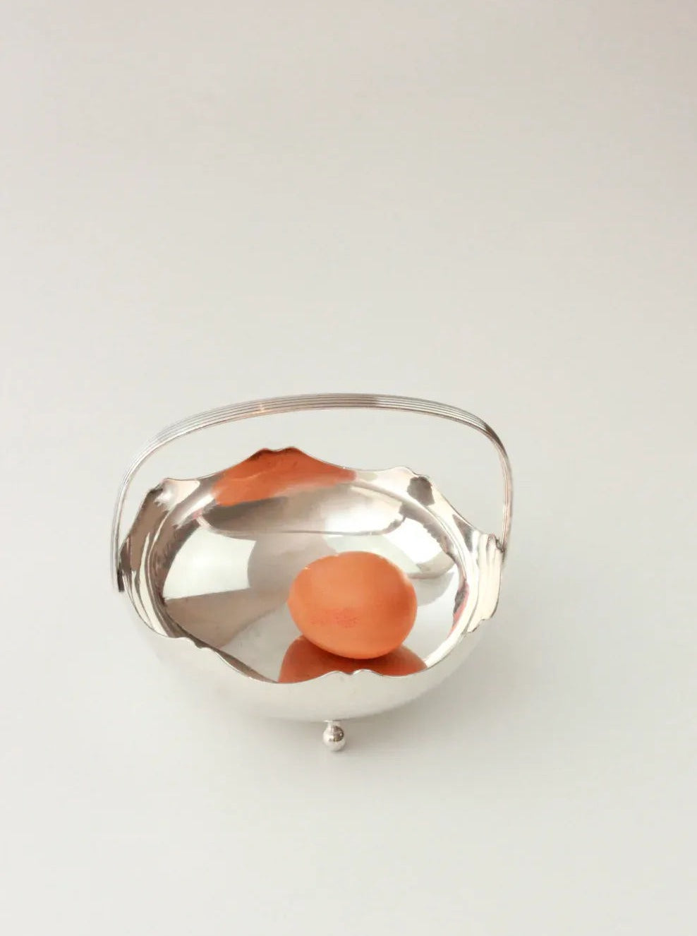 A single apricot is placed inside a Silver Vintage Basket by Boga Avante Shop. The basket, crafted in Meneses, Spain, features a shiny silver finish and a handle. It is set against a plain, light-colored background.
