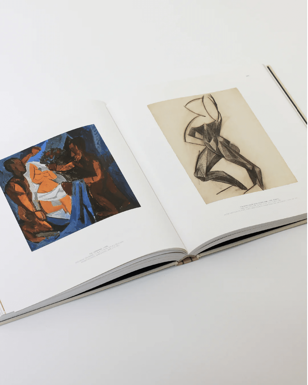 An open Cubist Picasso Book by Boga Avante Shop reveals two pages: on the left, a colorful abstract painting featuring figures reminiscent of Picasso's Cubist style; on the right, a monochromatic sketch showcasing geometric abstraction. The book lies elegantly on a light-colored surface, beckoning exploration of Cubism's influence.