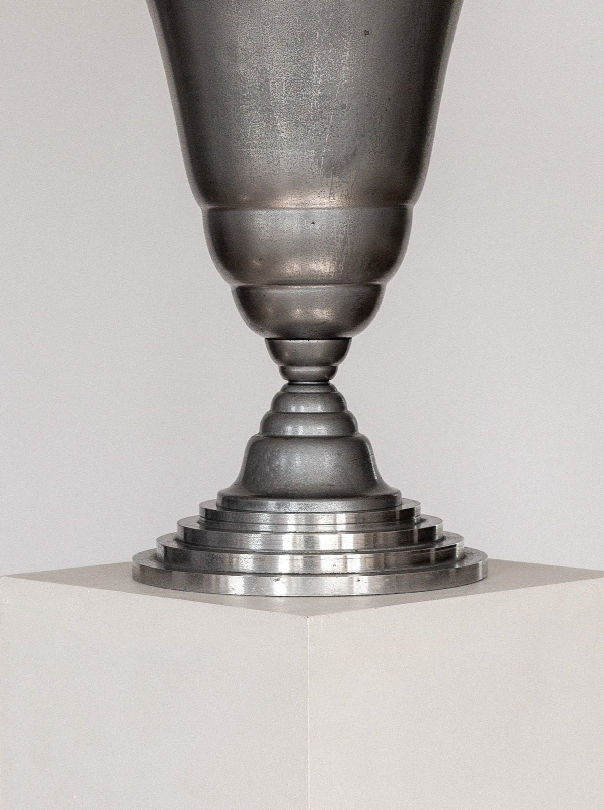A close-up of the Spigel Art Deco Torchière Table Lamp, France, displayed on a white pedestal. The oxidized aluminium lamp features a tapered design with an intricate, stacked base and a smooth, reflective surface. The background is a plain, light-colored wall that emphasizes the sculpture's details.