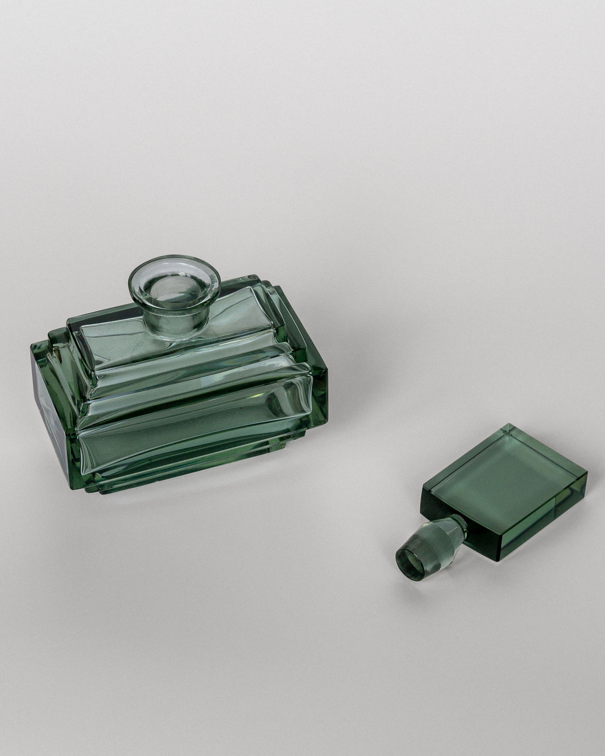 A Spigel Art Deco Perfume Bottle from the 1930s, featuring a rectangular, vintage-style green glass design, is shown lying on its side against a plain white background. The matching green glass stopper, also of Czech origin, is placed beside the elegant perfume bottle.