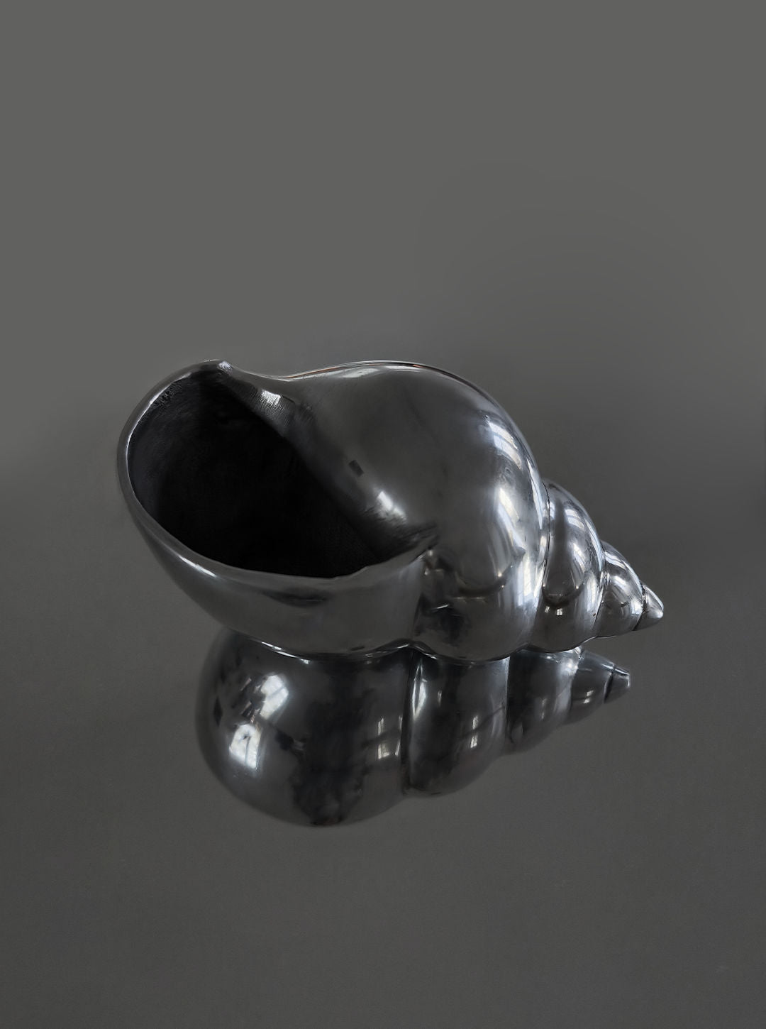 The Large Shell Sculpture by Les Objoies is displayed on a reflective surface, its glossy black spiral design mirrored below. A solid dark gray background enhances the sculpture’s lustrous texture.