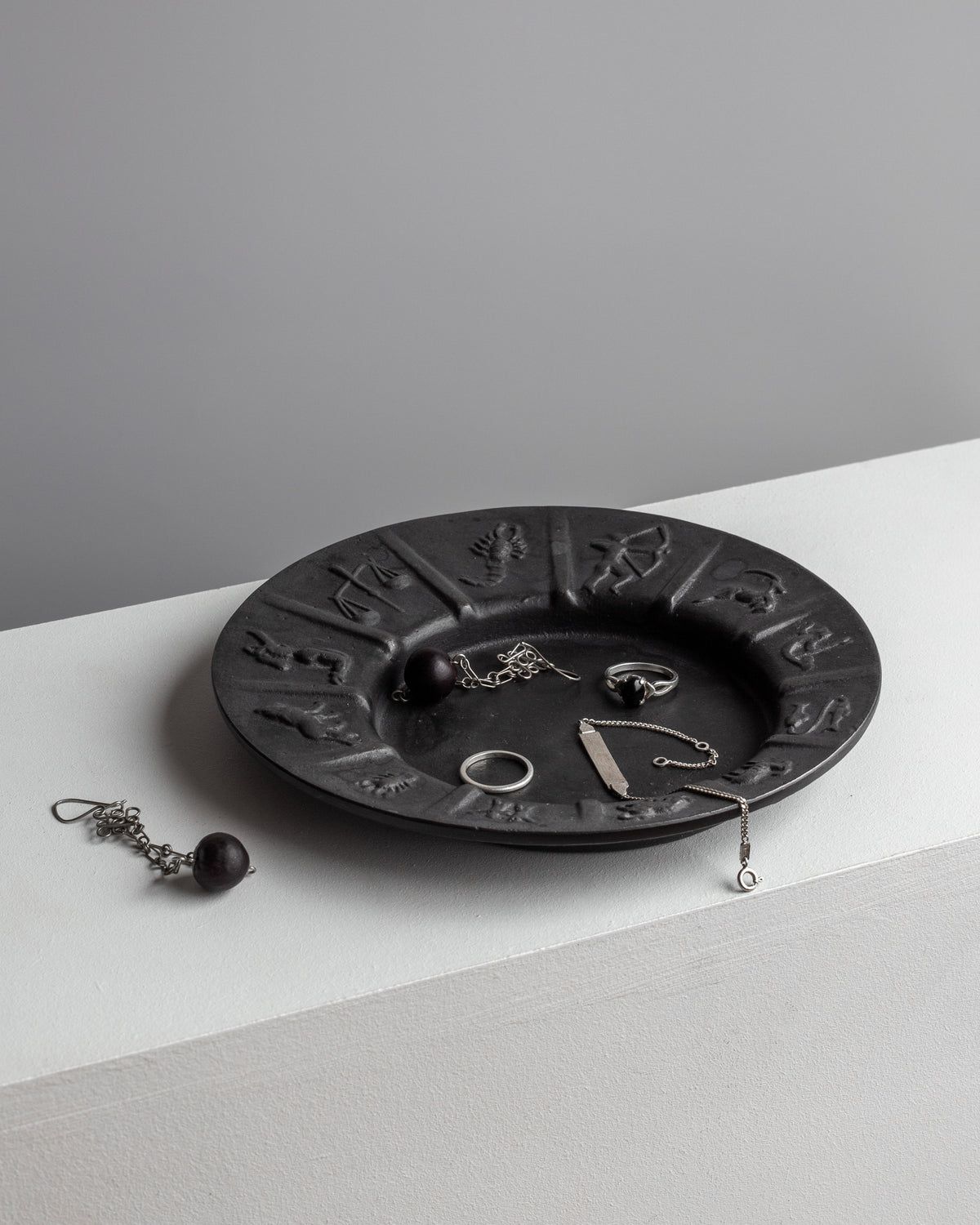 The Cast Iron Zodiac Dish by Spigel cradles an assortment of silver jewelry pieces, featuring a ring, necklace, and an earring with a dark bead, all displayed on a white surface against a gray backdrop.
