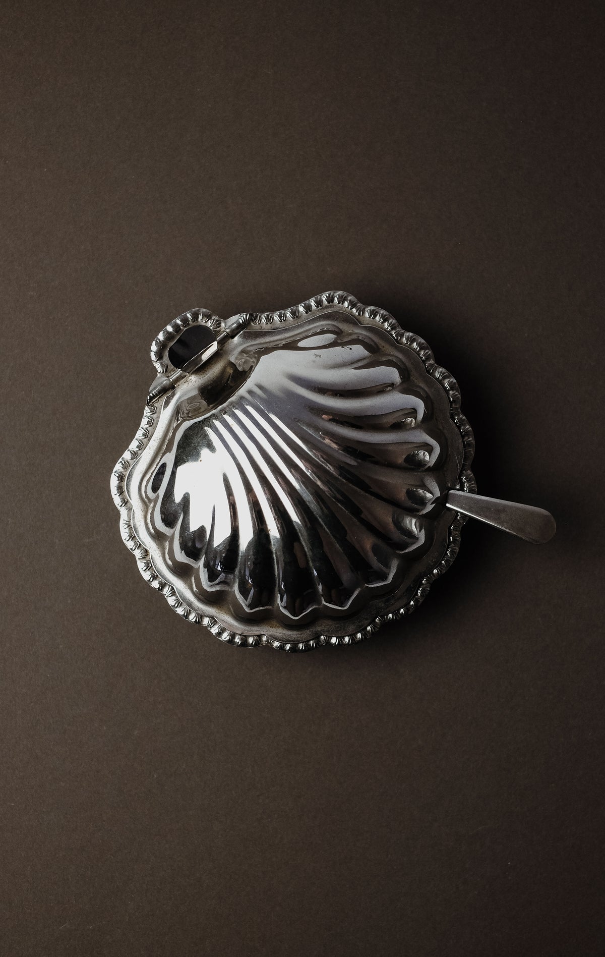 The Shell Shaped Butter Dish with Spoon by septembre studios is an elegant addition to your dining experience. This polished silver dish, with a lid resembling a seashell and adorned with decorative ridges and an ornate handle, rests stylishly on a dark matte surface.