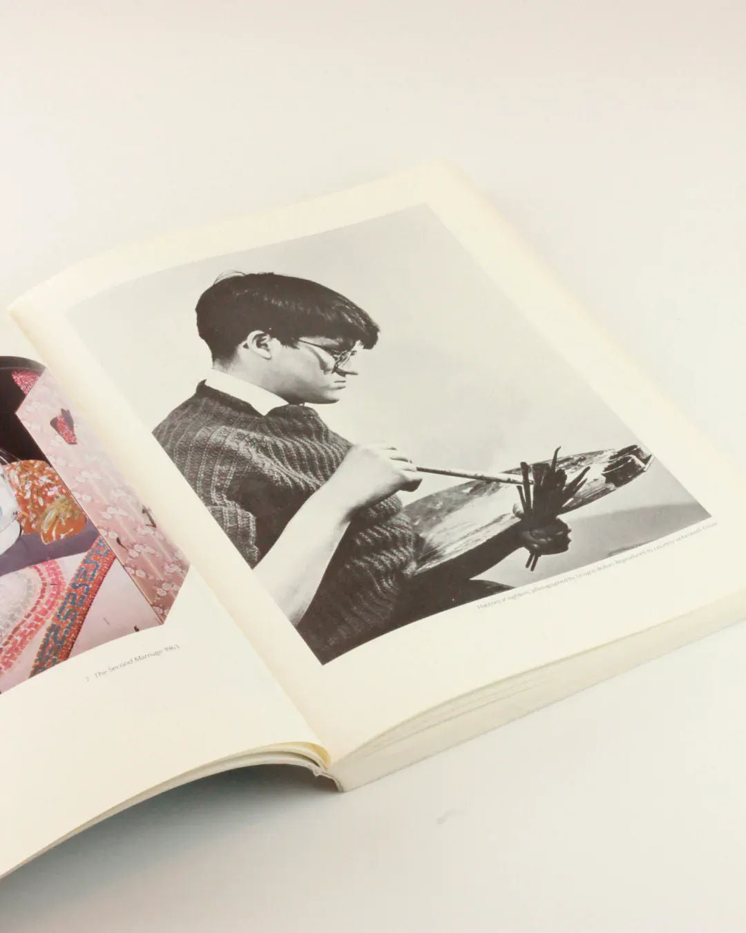 An open David Hockney 'My Early Years' from Boga Avante Shop reveals a black-and-white photograph of a person knitting. The individual, wearing glasses and a sweater, is deeply focused on their craft. Adjacent is a partially visible colorful image, evoking the vibrant style of David Hockney in the context of arte británico.