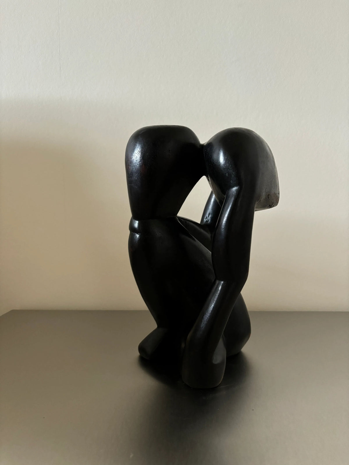 A CU-RA-TED Black Wooden Object - "The Thinker," of an abstract human figure bending forward with one arm extended downwards and the other reaching slightly backward. The figure's head is smooth and featureless, showcasing exquisite craftsmanship. The Black Wooden Object rests on a simple, flat surface against a plain background.