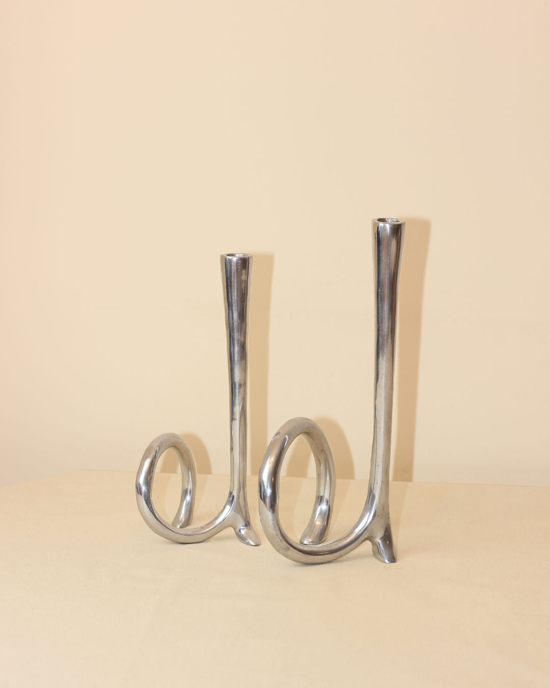 The Cast Aluminum Candleholders by Boga Avante Shop feature abstract, looped silver bases inspired by 1980s candelabras—ideal for modern minimalist decor, displayed on a beige surface with a light beige background.