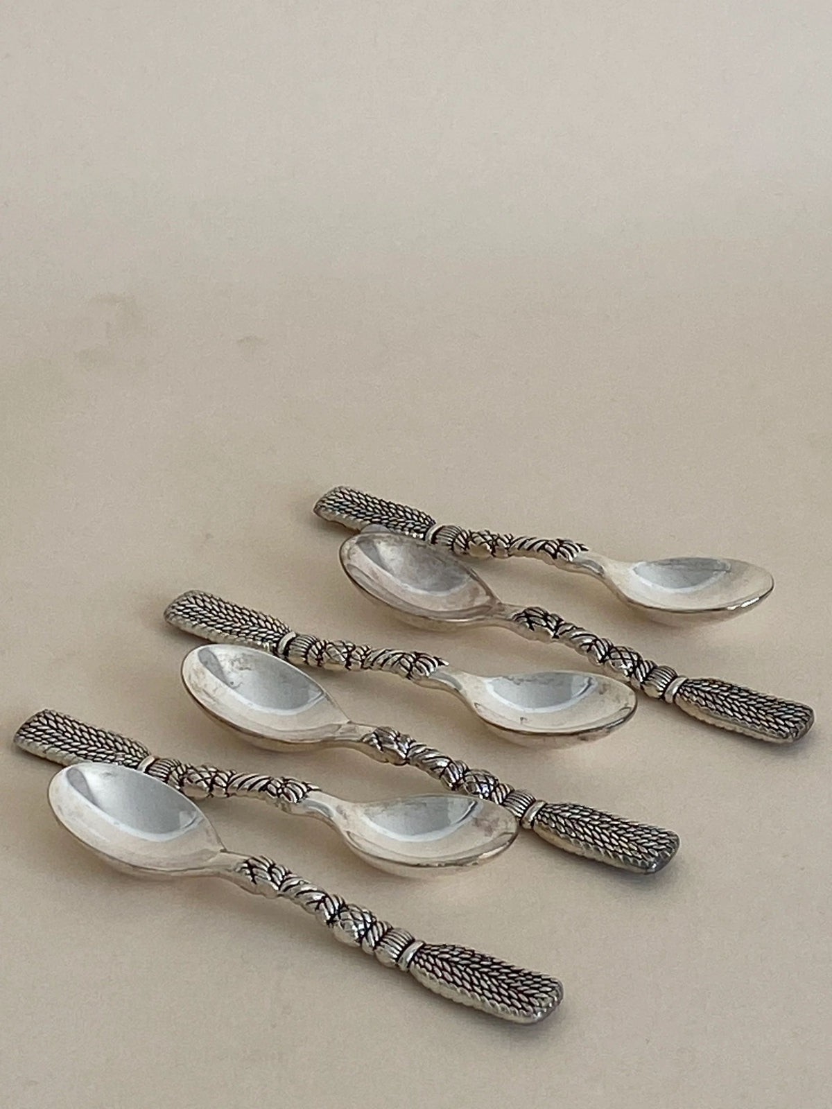 Five Aesthete Label French Tassel Spoons with intricately designed, textured handles are arranged casually on a light beige surface. The silver-plated spoons, in excellent vintage condition, feature a unique pattern resembling braided or woven metal. Reflecting light softly, they suggest a polished finish—perfect for a decorative table setting.