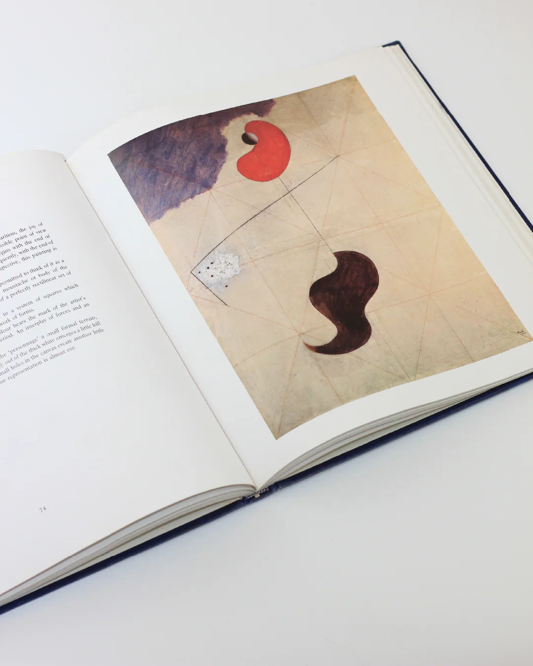 The Mirò The Masterworks Book from Boga Avante Shop rests open on a white surface, showcasing a surrealist modern art piece with red and brown shapes on a beige background, divided by thin geometric lines. The opposite page contains text highlighting Joan Miró's imaginative essence.