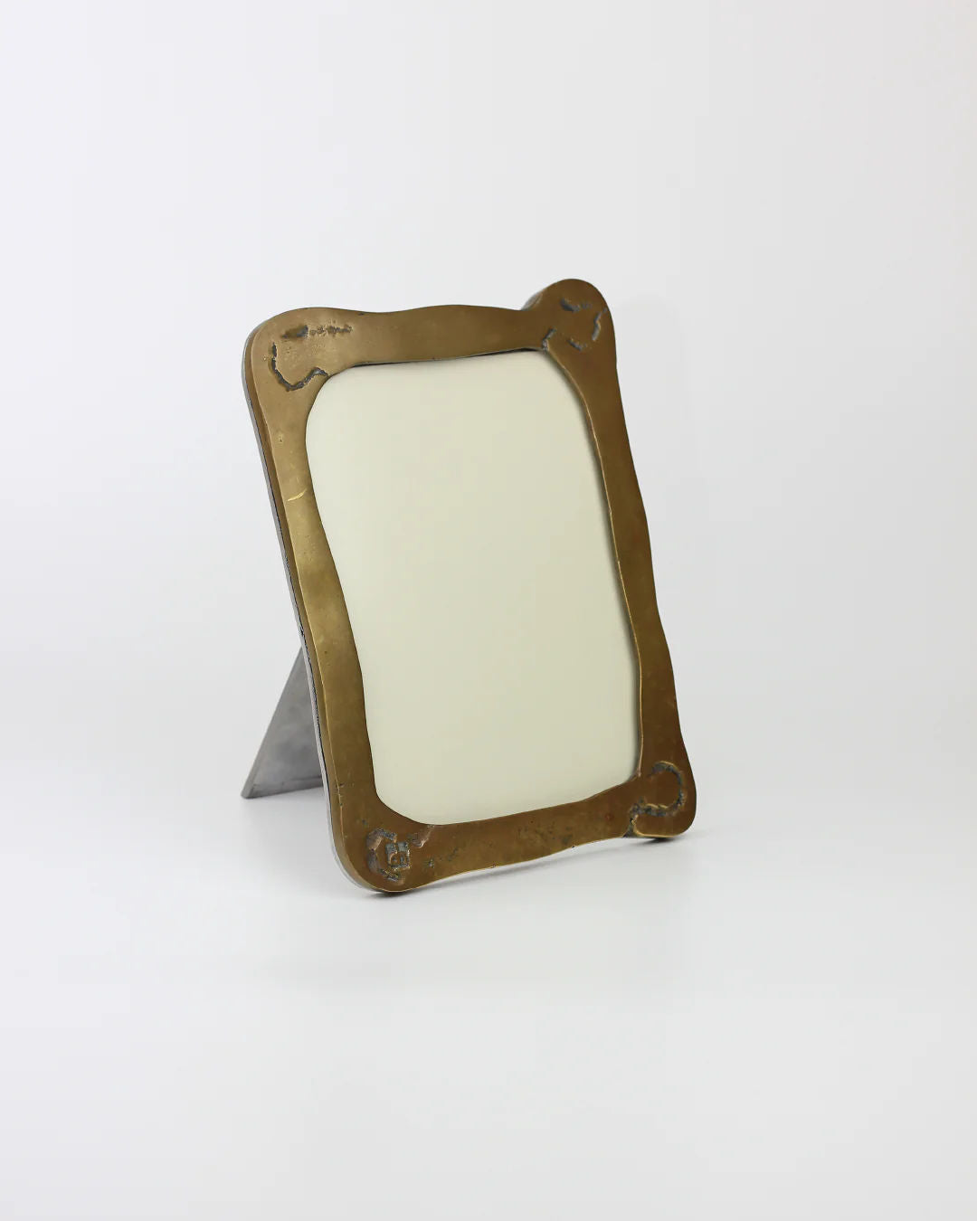On a white surface stands the Brutalist Mirror by Boga Avante Shop. This minimalist bronze frame with empty white space has rounded, ornate edges for a subtle industrial finish and includes a stand at the back for upright positioning.