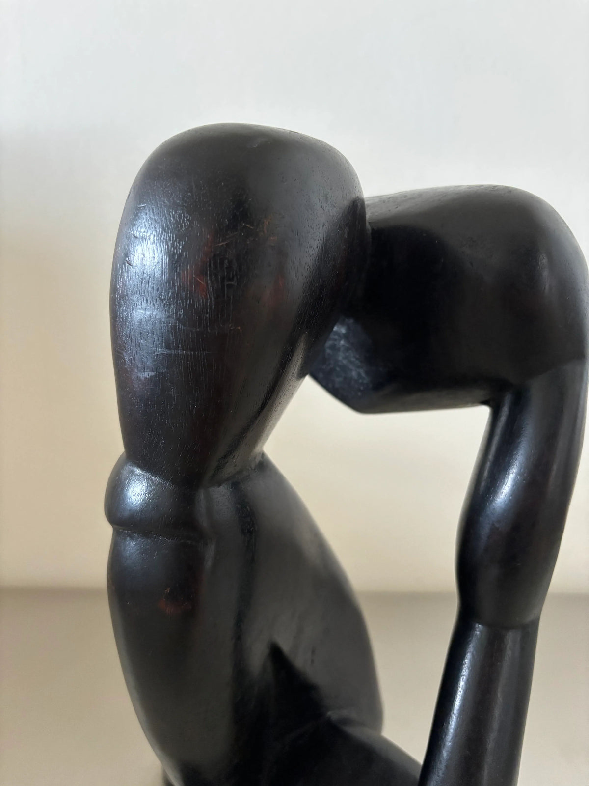A close-up photo of an abstract, smooth Black Wooden Object - “The Thinker” by CU-RA-TED shaped in a seated, contemplative pose with its elbow resting on its knee and hand under its chin. The artistry showcases impeccable craftsmanship with a minimalist design, lacking facial features or detailed anatomy.