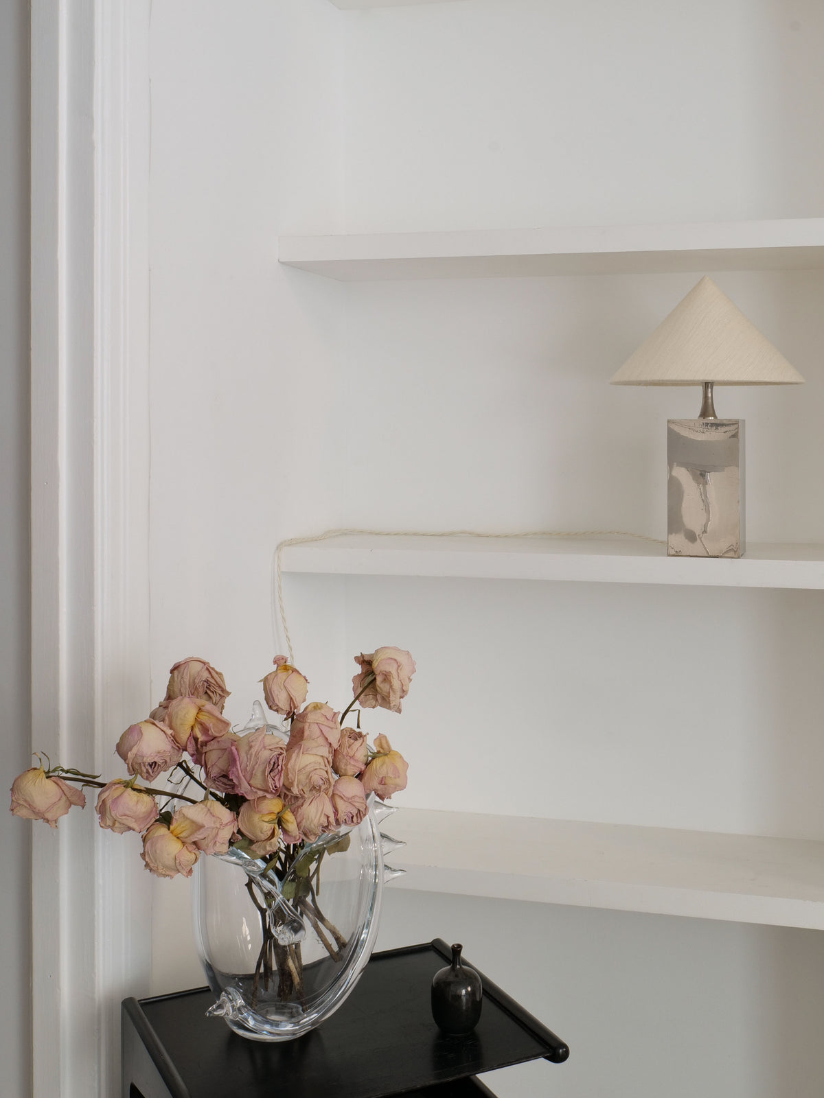 A minimalist shelf set against a white wall holds a Barbier Small Box Lamp 1970s by Collection apart, featuring a beige shade and rectangular base. Below, a black table supports a transparent glass vase containing wilted pink roses and a small dark ornament, evoking the elegance of French 1970s lighting.
