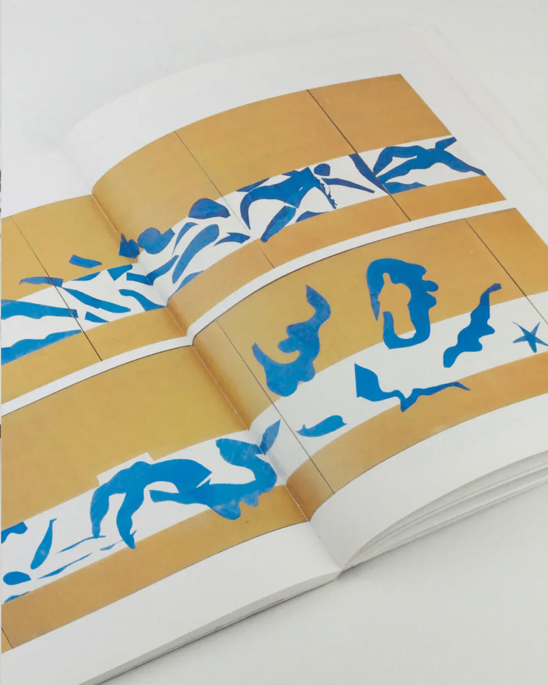 In the open book "The Cut Outs of Henri Matisse" by Boga Avante Shop, multiple panels showcase a series of abstract blue shapes set against a yellow background. These patterns, reminiscent of Matisse's mature cut-outs, feature fluid, organic forms with sweeping curves and sharp edges. The artwork spans two facing pages, creating a seamless visual flow.