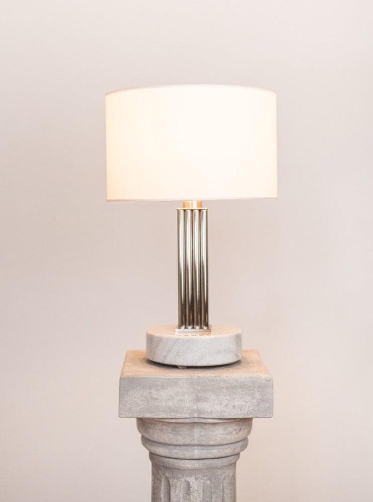 The Athens table lamp 4 by Brajak Vitberg, featuring a cylindrical white shade and sleek metallic base, sits on a stone pedestal. The softly lit background enhances its sculptural elegance, making it an outstanding piece for luxury interiors.