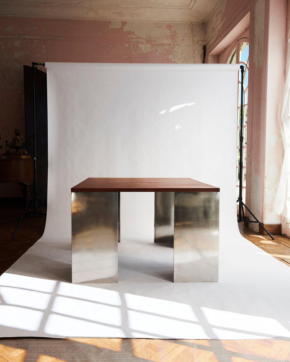 The Square Table 2022 by Studio HAOS, featuring a wooden top and reflective metal legs, stands before a white backdrop in sunlight. Pink walls, arched windows, and soft shadows create grounded elegance in the serene space.
