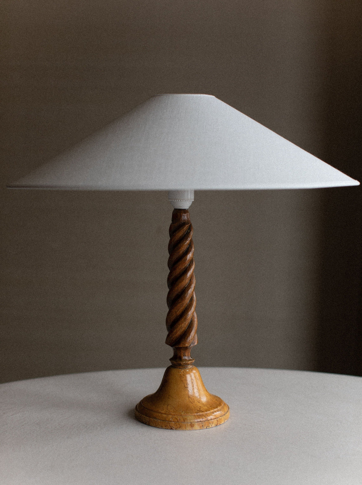 A Twisted Wooden Lamp by Out For Lunch with a wide, white conical lampshade and a twisted base stands on a round white table. The natural wood grain showcases intricate spiral carvings, highlighting its artisanal craftsmanship. The background is a neutral, blurred wall.