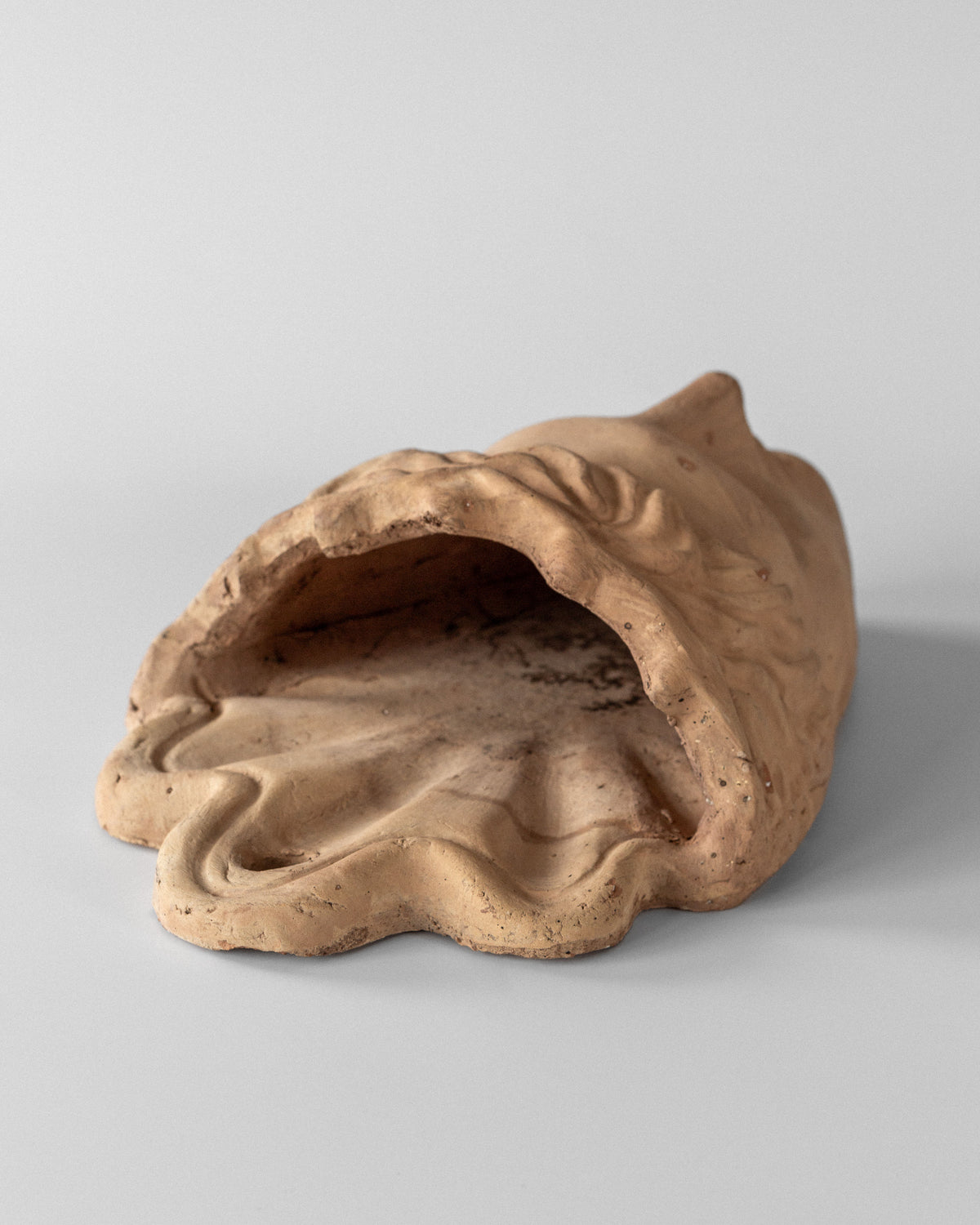 The Spigel Terracotta Wall Mask Planter resembles a draped, hollow form lying on its side against a gray background. Its rough texture features subtle color variations and surface details reminiscent of neo-classical design.
